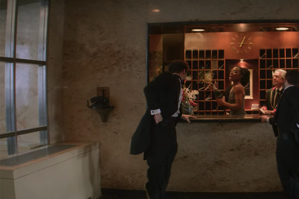 Andrew Garfield and Robin de Jesus dance in coats and tails in front of a concierge window where Luis Miranda, Jr. plays a concierge. 