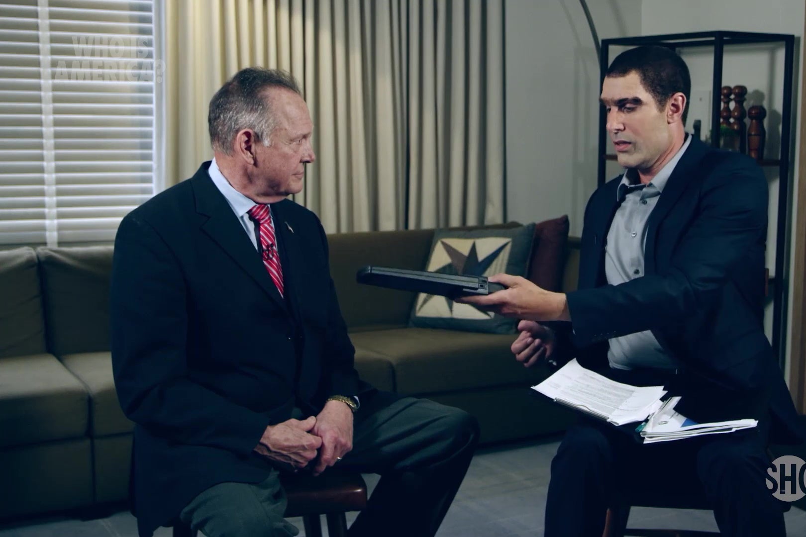 Roy Moore sits in a chair while Sacha Baron Cohen, wearing a ridiculous, lantern-jawed disguise, waves a metal-detector over him.