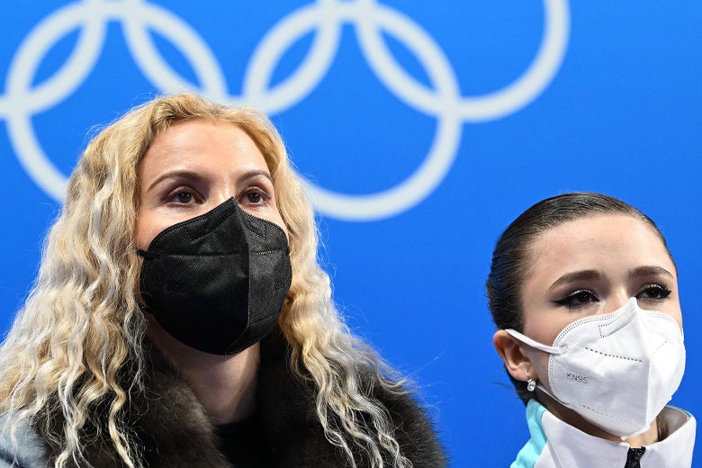 Tutberidze and Valieva in KN95 masks looking upward with the Olympic rings on the backdrop behind them.