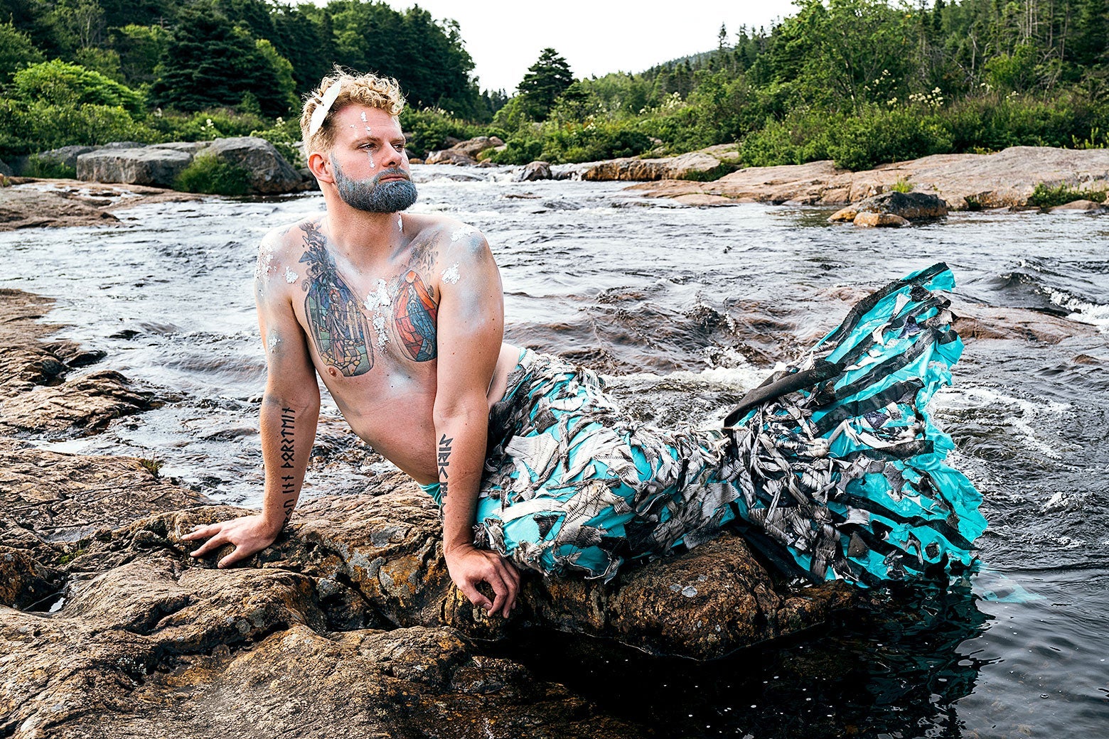 A merman with a blue tail and glittery makeup basks on a rock. 
