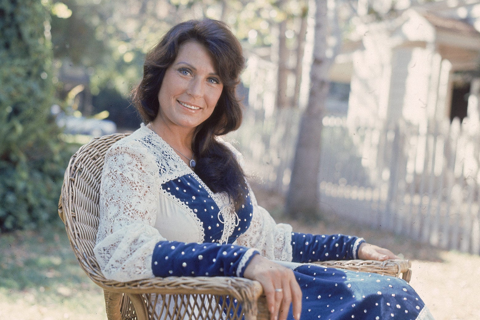A portrait of shows the singer and guitarist sitting outside in a wicker chair, wearing a blue-and-white lacy dress, and long faintly curly brown hair, smiling at the camera