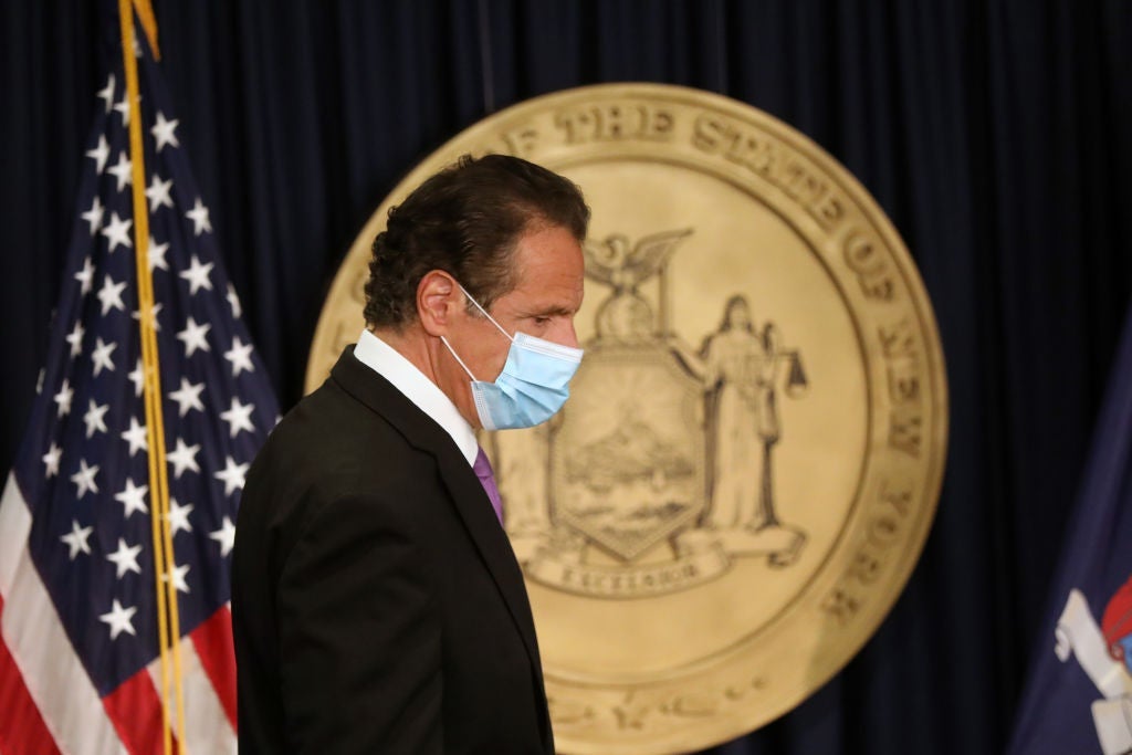 Cuomo, wearing a mask, walks in front of a state of New York seal.