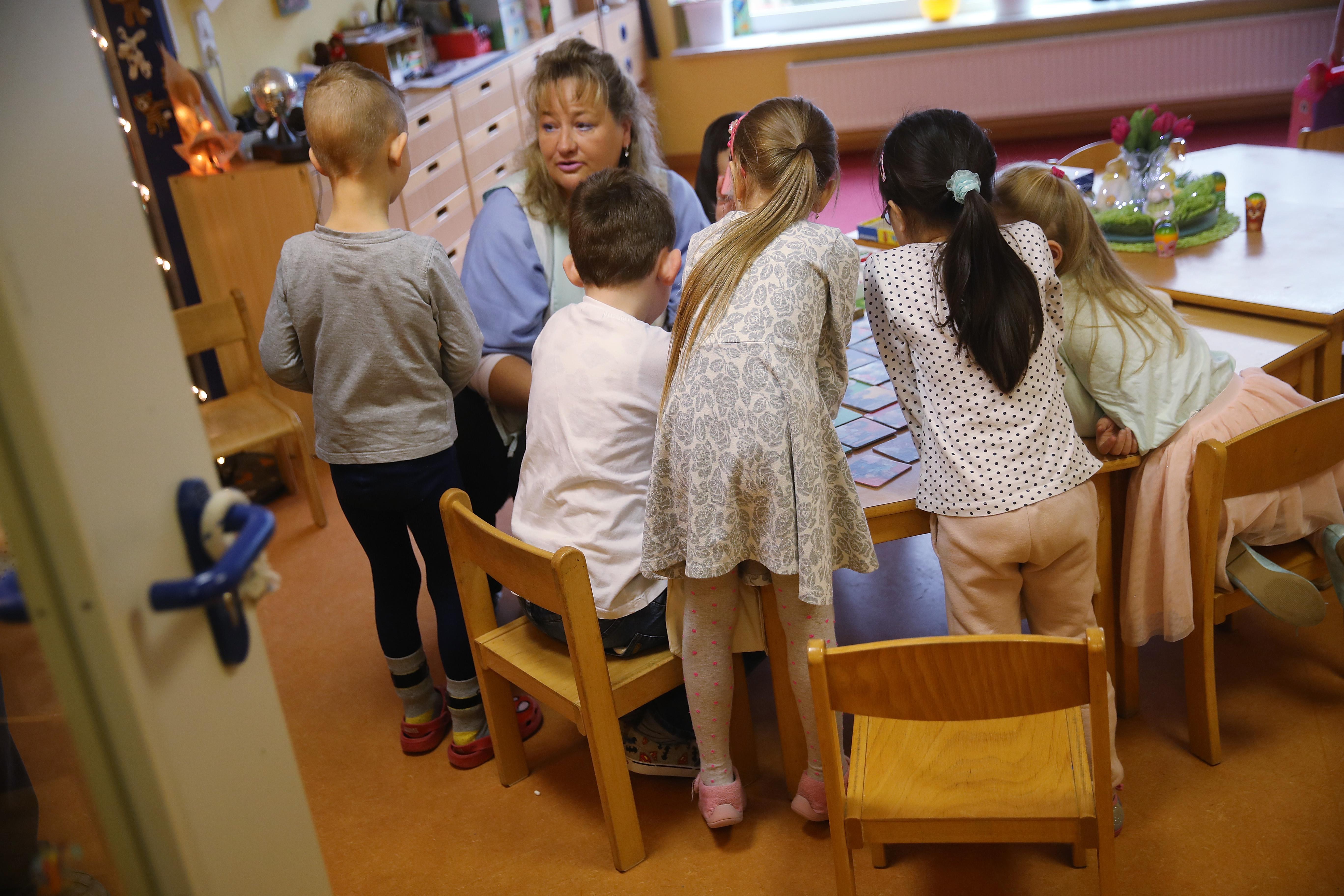 A nursery school teacher supervises children in a day care center with tables and toys.