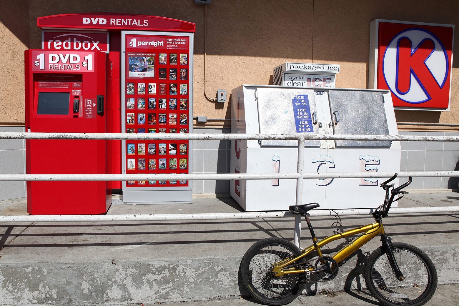 A RedBox video rental kiosk in front of a gas station. A yellow bike is parked nearby.