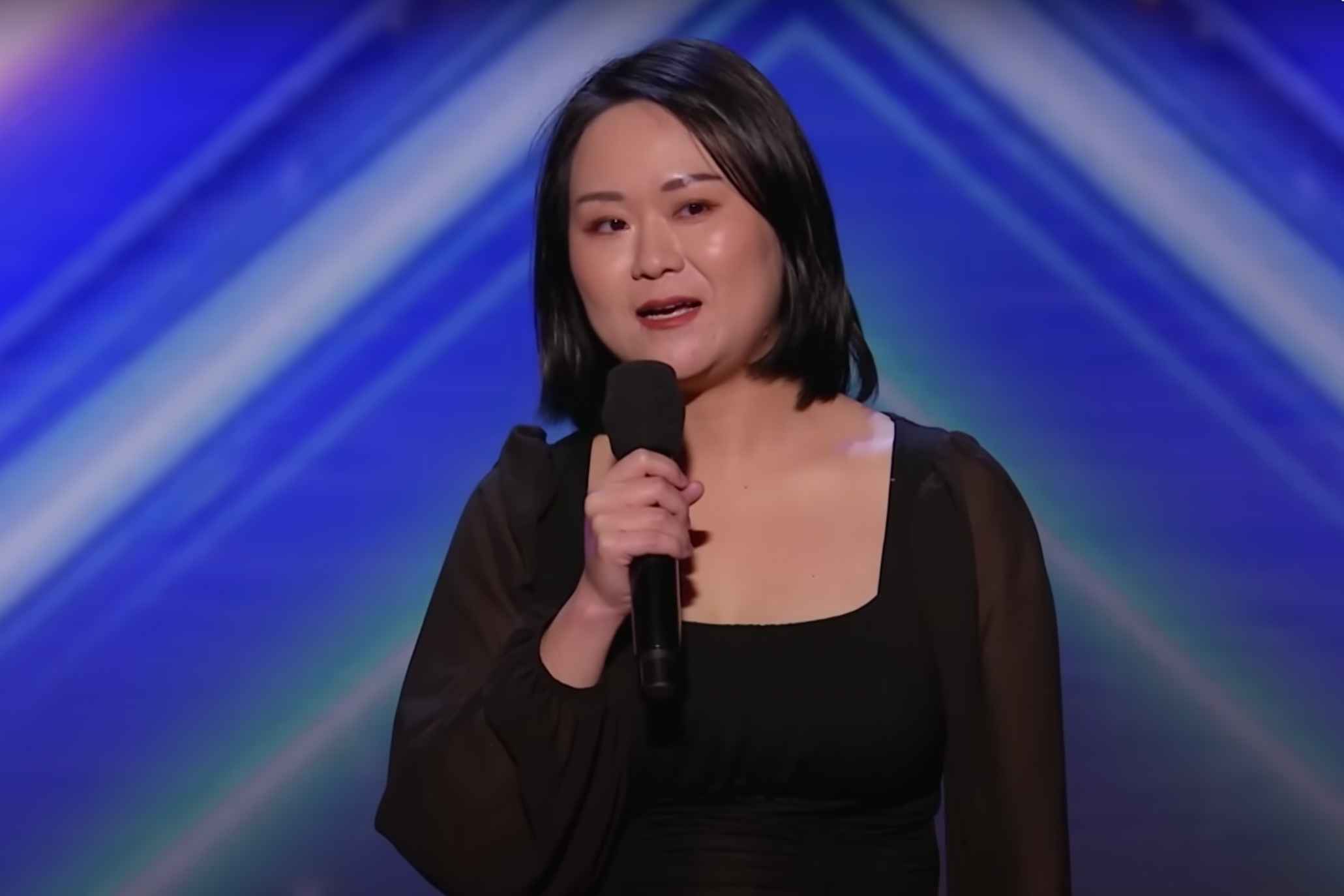 She wears straight black hair down to her shoulders and a simple black blouse, a microphone in her hand and a sparkly blue backdrop behind her