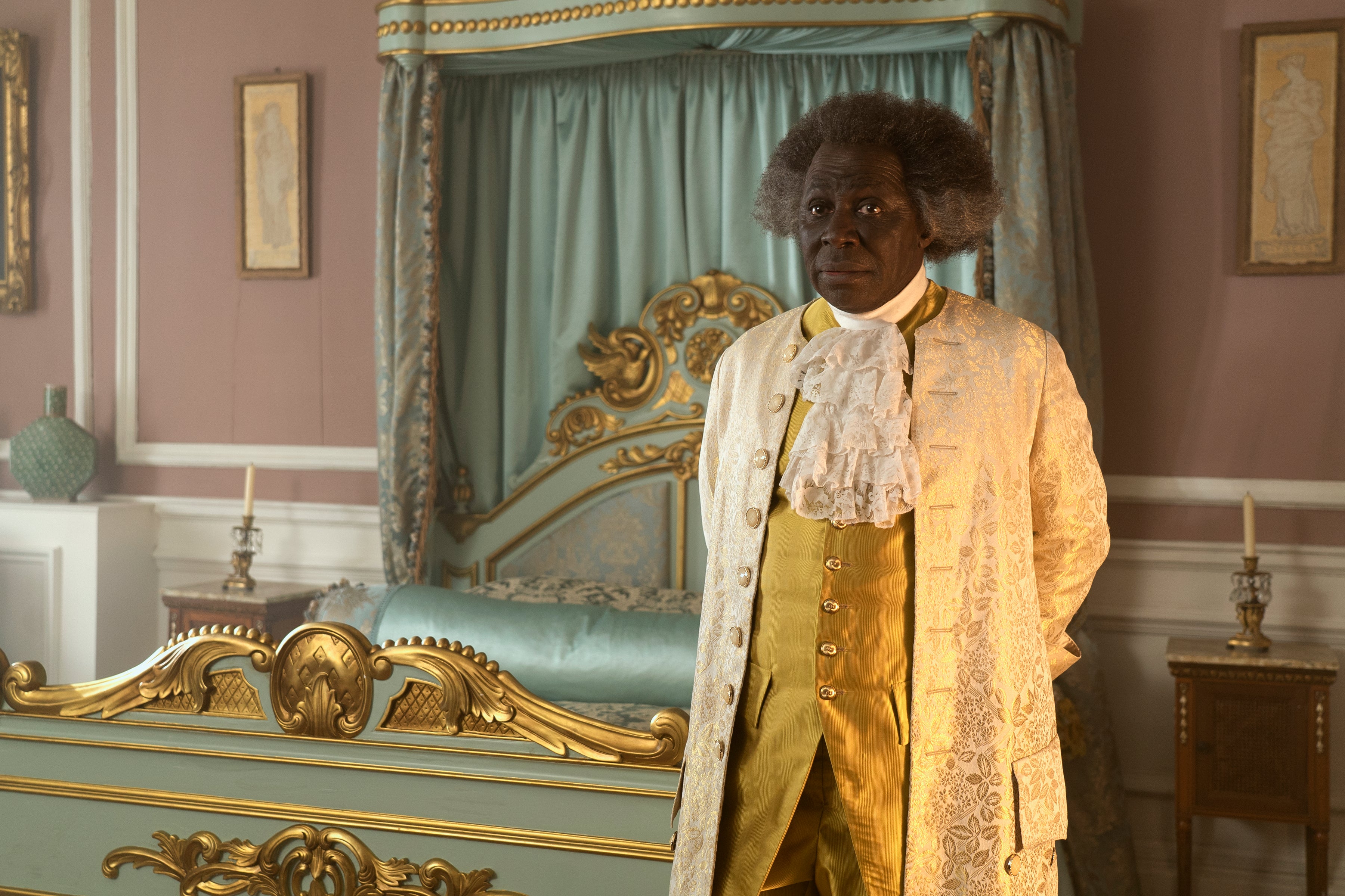 A Black man with gray hair stands next to a bed and wears a gold-colored Georgian suit.