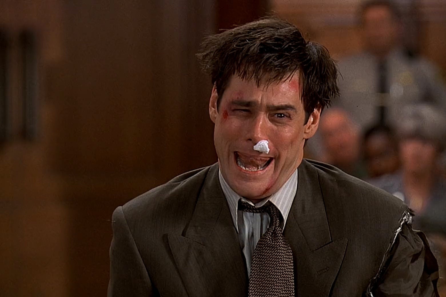 Jim Carrey faces the camera in a courtroom with a torn suit jacket, askew tie, piece of cotton in his nose, blood and bruises on his face, and mussed hair. He stands with his mouth open. 