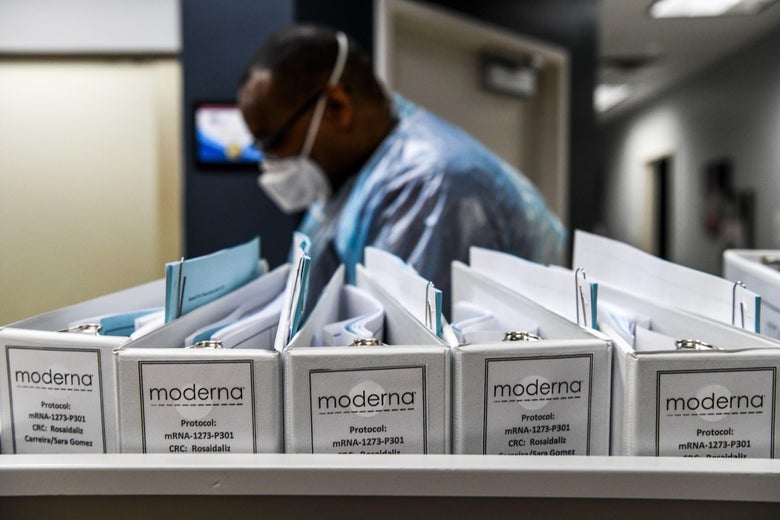 Folders labeled "Moderna" are seen in front of a person wearing PPE.