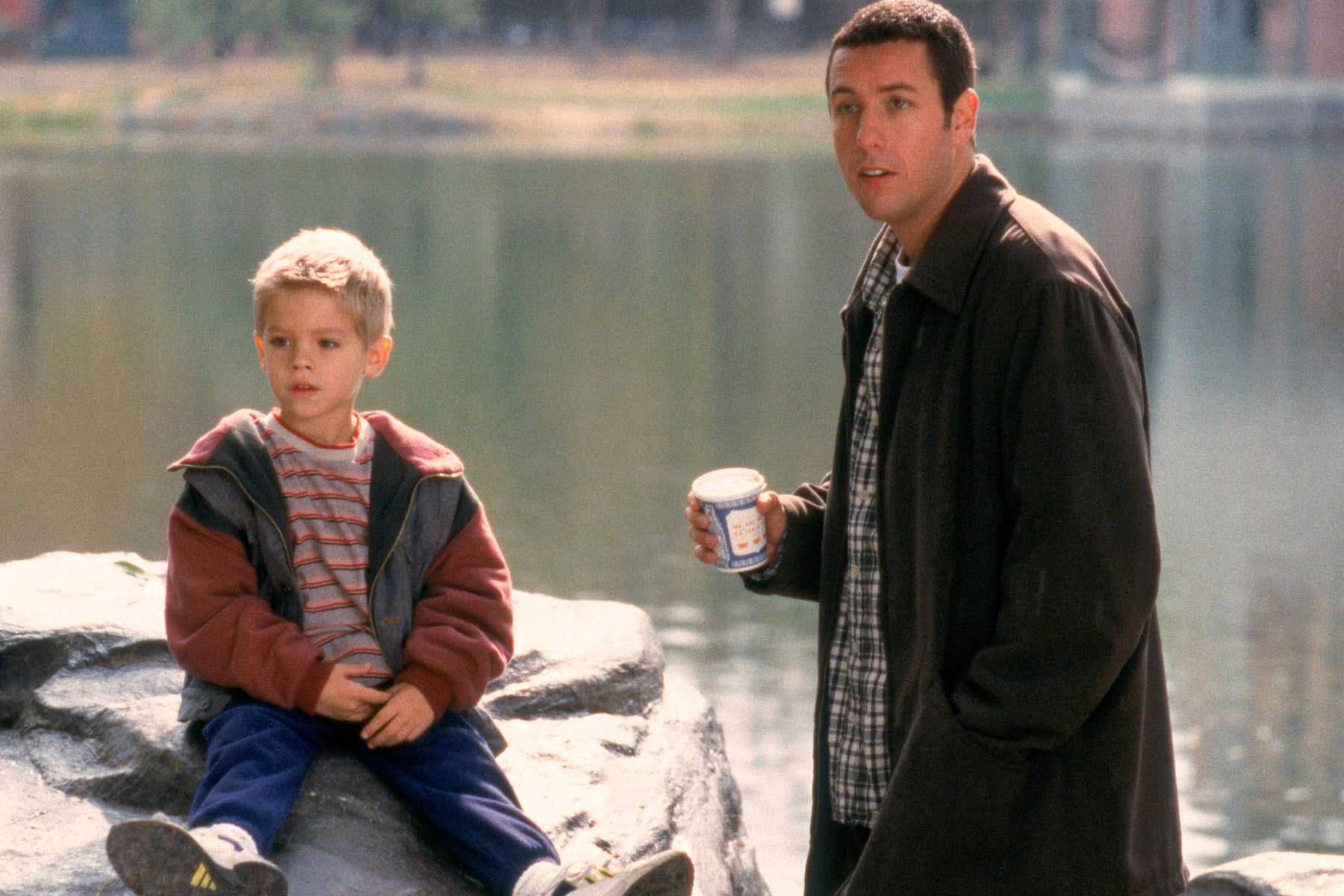 A young Cole Sprouse sits on a boulder looking off camera. Adam Sandler stands beside him, holding a coffee cup, and looking in the same direction. 