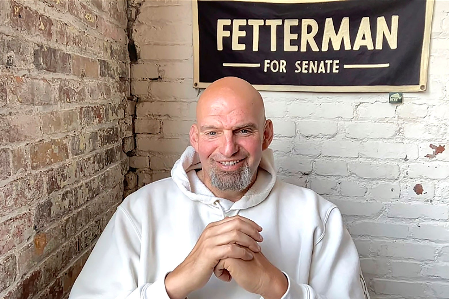 John Fetterman speaks during a video interview from his home wearing a baggy white hoodie and a banner bearing his name behind him.