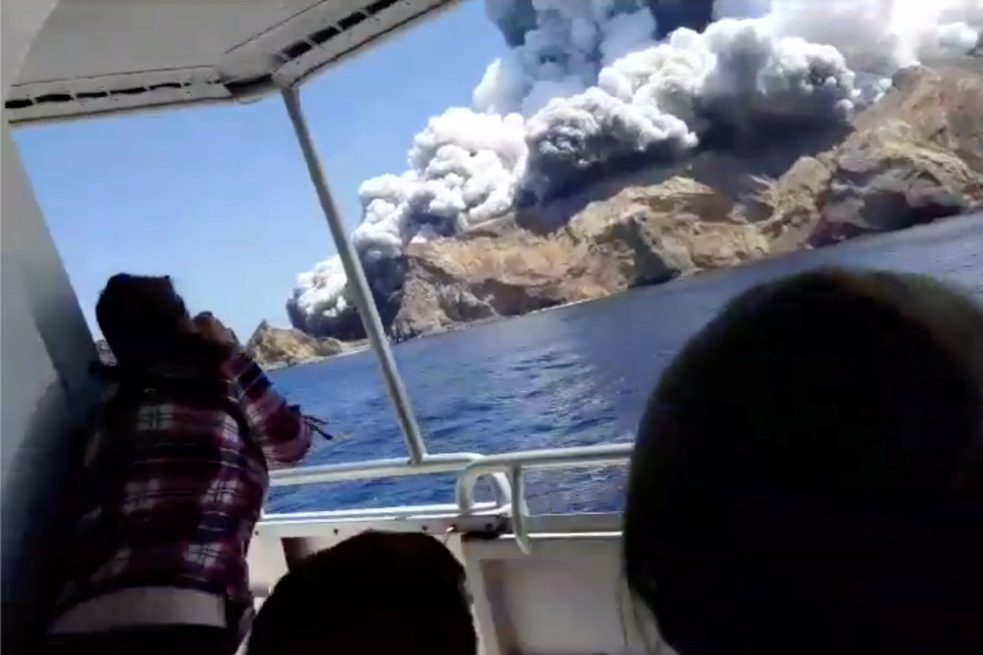 The volcanic eruption is seen from inside a boat. A passenger in the boat leans forward, possibly to take a photo.