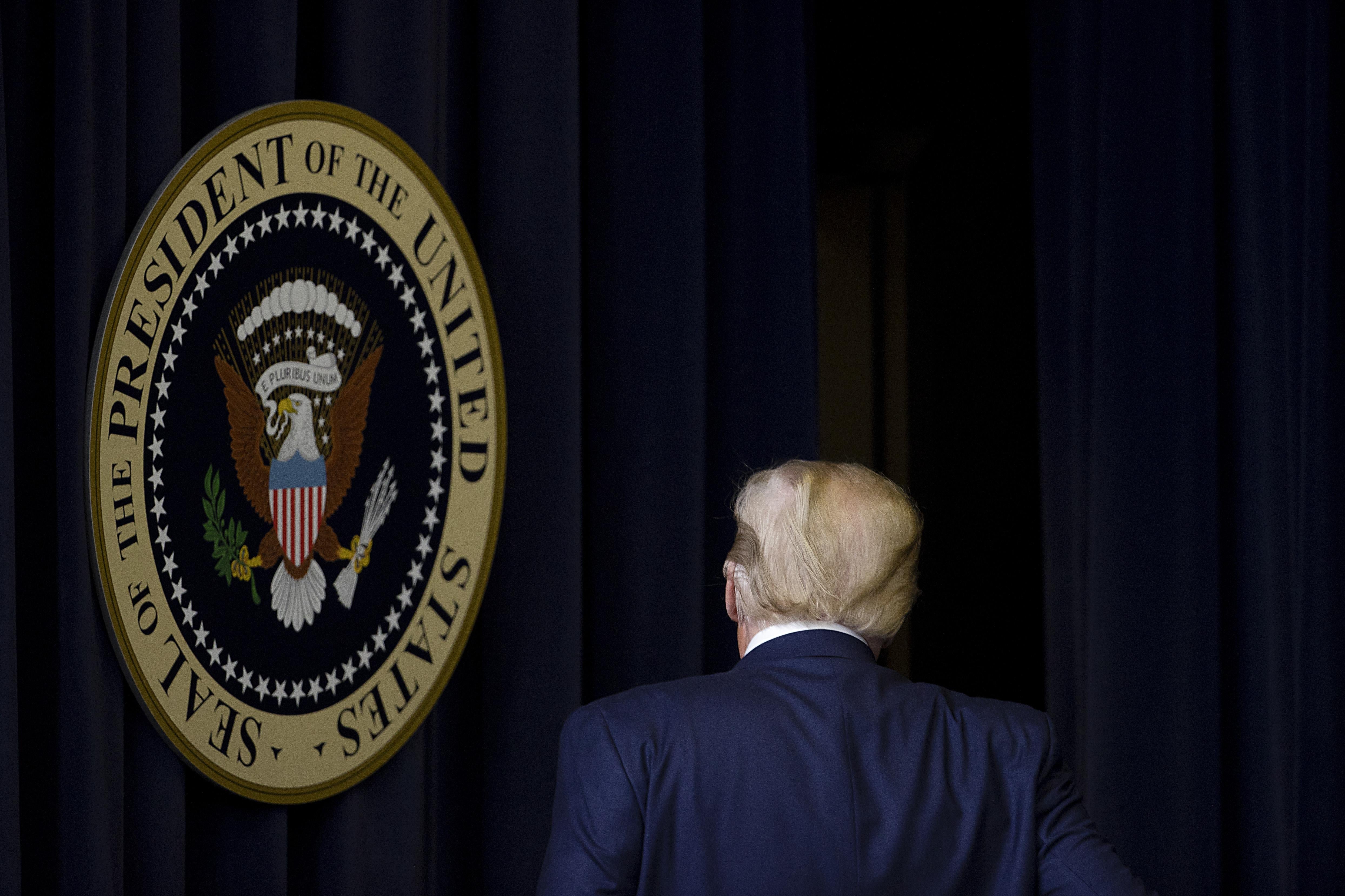 Trump, seen from the back, walking past the presidential seal hanging in front of a curtain