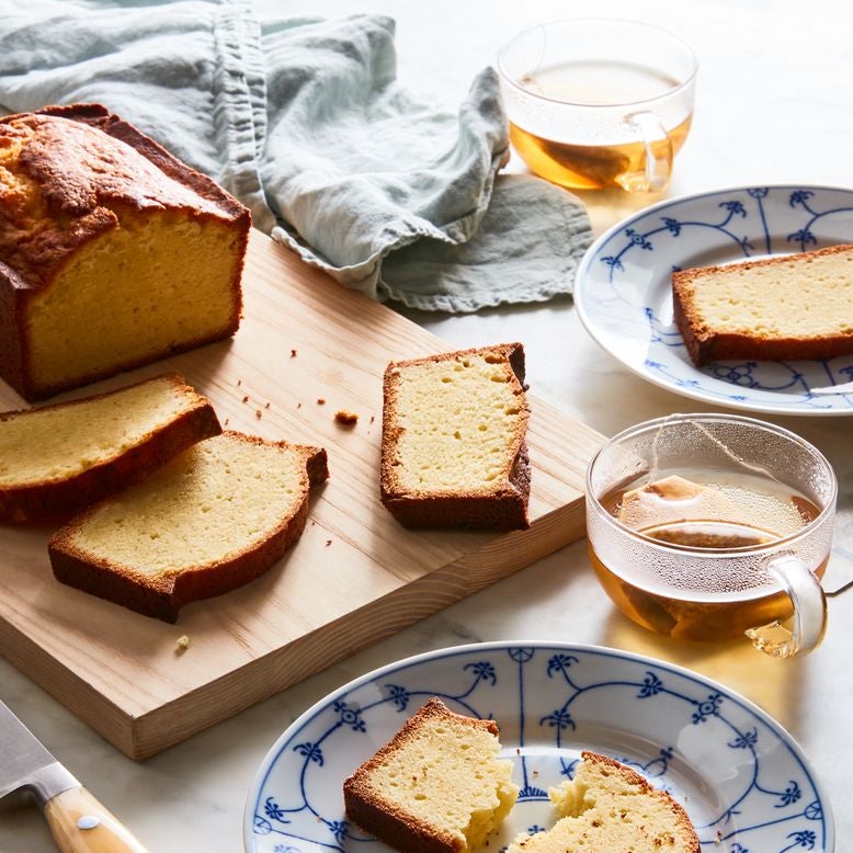 A loaf of plain pound cake sliced on a wooden cutting board, surrounded by individual slices of pound cake on small plates, two glass cups of tea, and a knife.