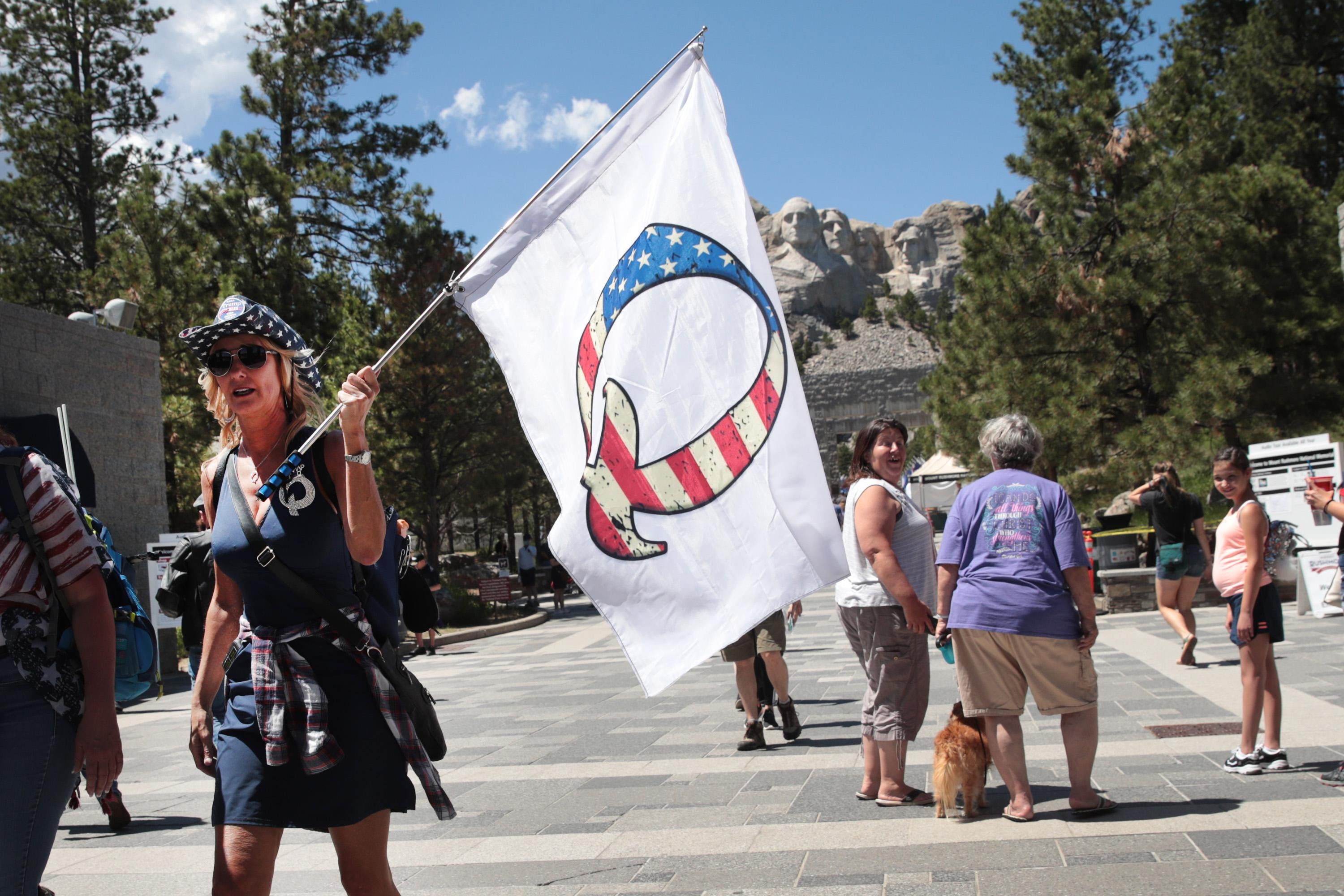 A person wearing blue and a cowboy hat walks while holding a white flag with the letter "Q." In the background Mount Rushmore is visible.