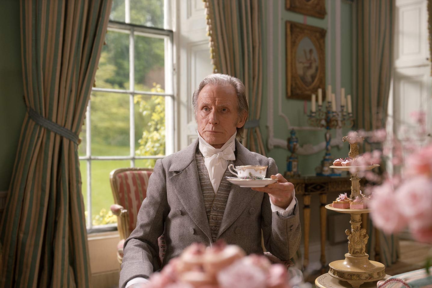 Bill Nighy sits in a lavishly furnished room holding a cup of tea in a saucer. A nearby table is covered in towers of pink desserts.