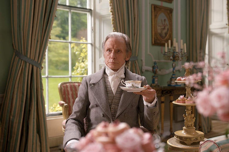 Bill Nighy sits in a lavishly furnished room holding a cup of tea in a saucer. A nearby table is covered in towers of pink desserts.