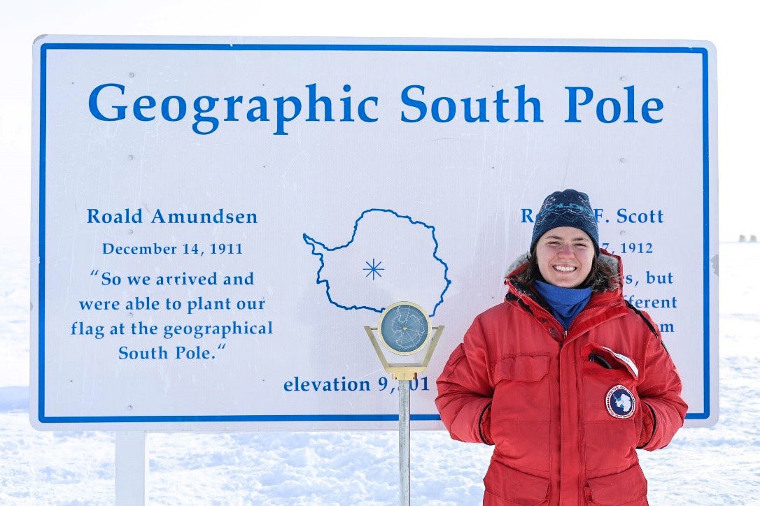A young woman smiles next to a sign that says "Geographic South Pole."