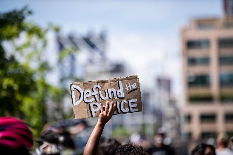 A hand is seen holding up a sign that says "Defund the Police."