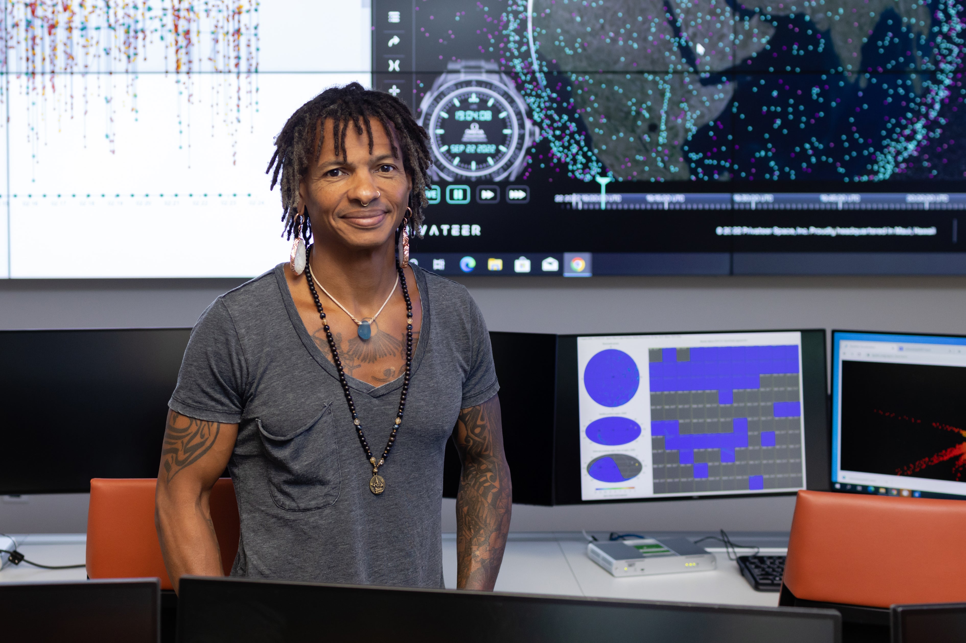A man with dreadlocks and earrings stands amidst an array of computers and video screens showing sciencey stuff.