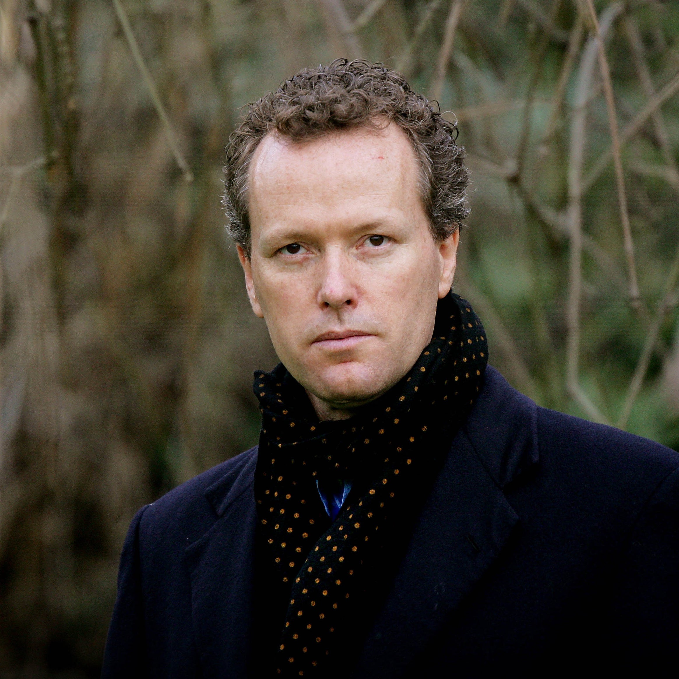 A man in an overcoat stands in front of trees.