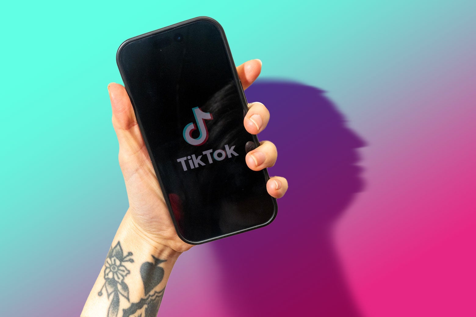 Hand holding a phone showing the TikTok logo. There are black tattoos on the person's wrist. The phone is casting a shadow of Donald Trump's face and the background color is a gradient of blue, purple, and pink.