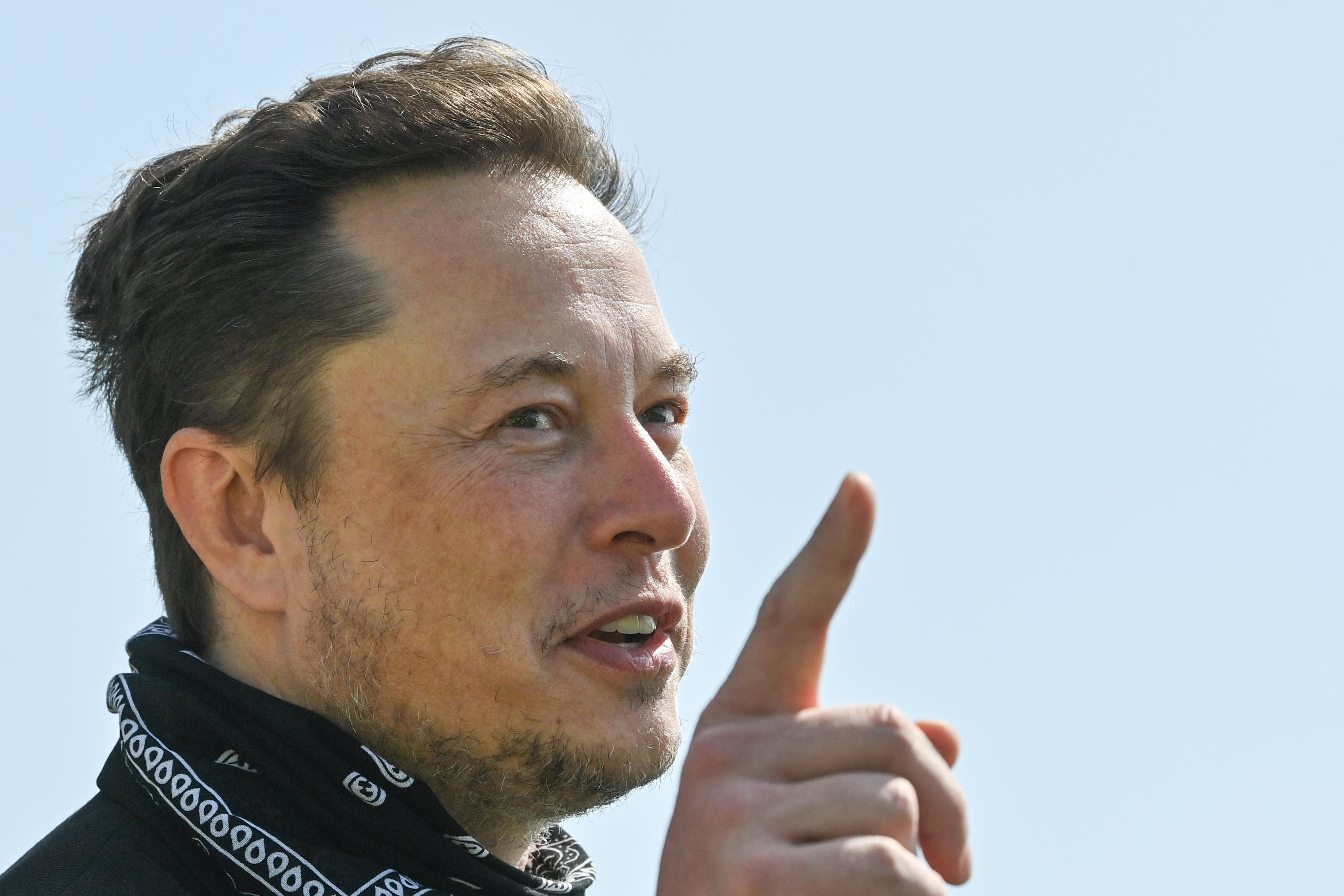 Musk pointing and smiling as he speaks outside against a blue sky