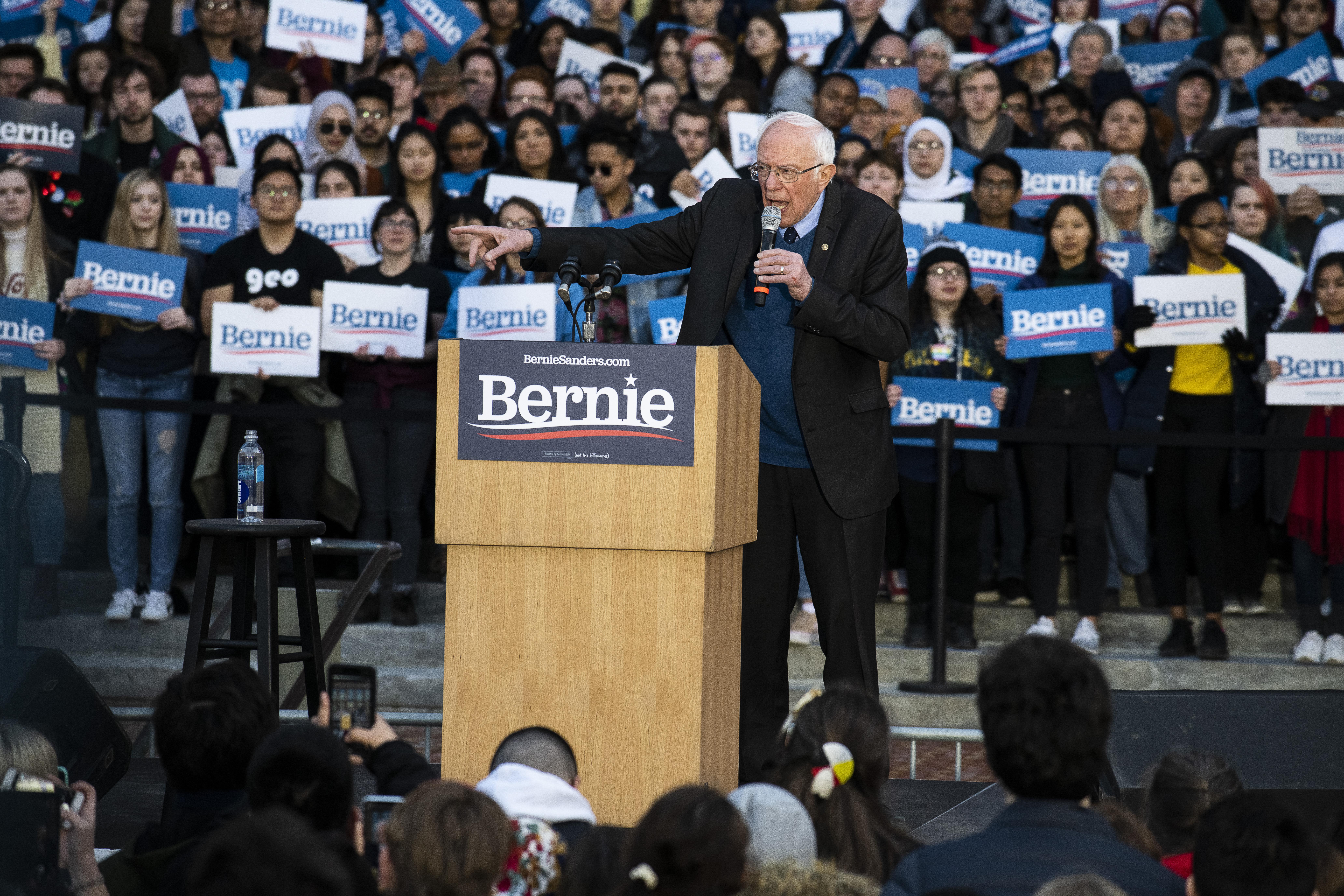 Bernie Sanders speaks at a podium with a crowd of supporters surrounding him.