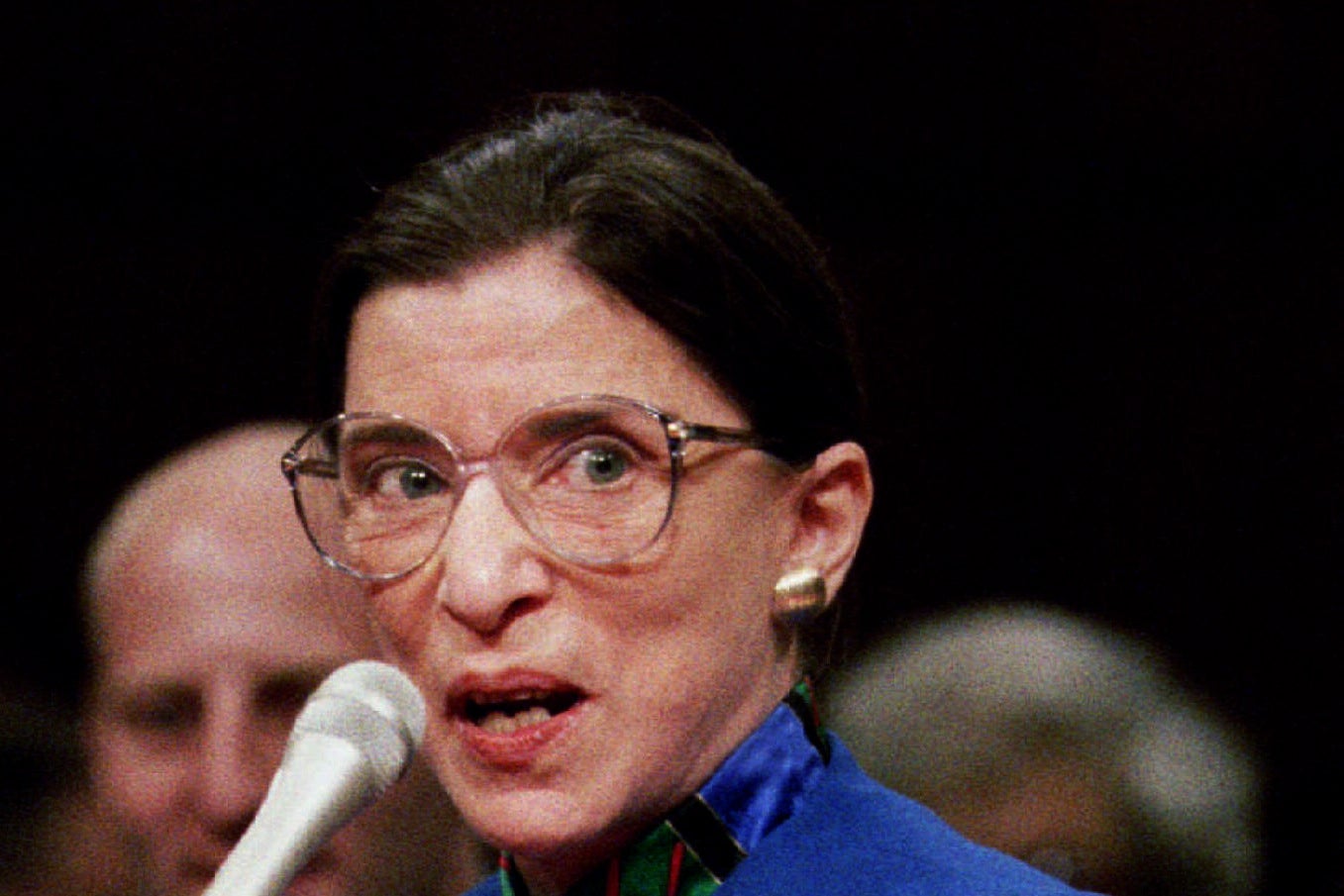 Ruth Bader Ginsburg behind a microphone in big glasses and a blue jacket.