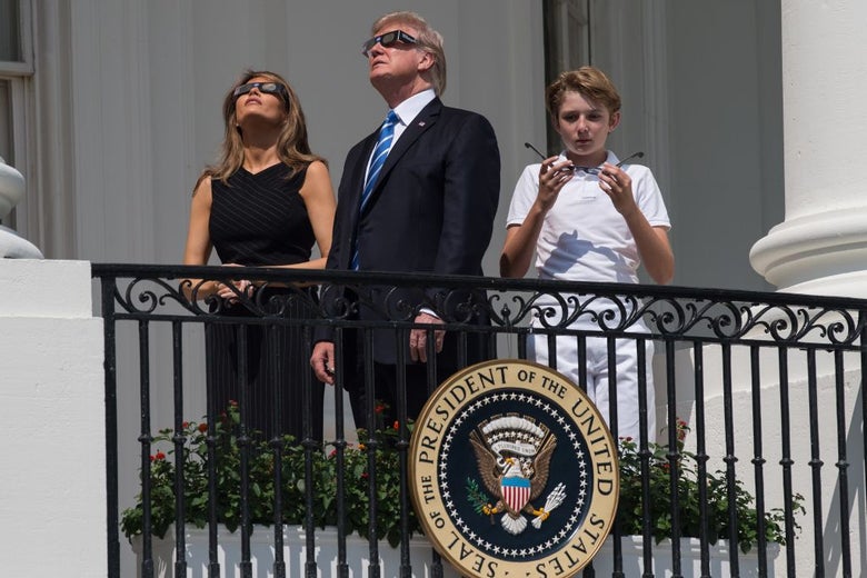 The family stands on a balcony whose wrought iron railing holds up a large presidential seal. Barron is looking at his eclipse glasses and Melania is looking into the sky. Donald is turned slightly to the side, looking upward, but seemingly at something distinct from what Melania is looking at.