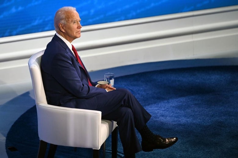 Biden, seated in a white chair on a blue carpeted stage, looks up and to his right.