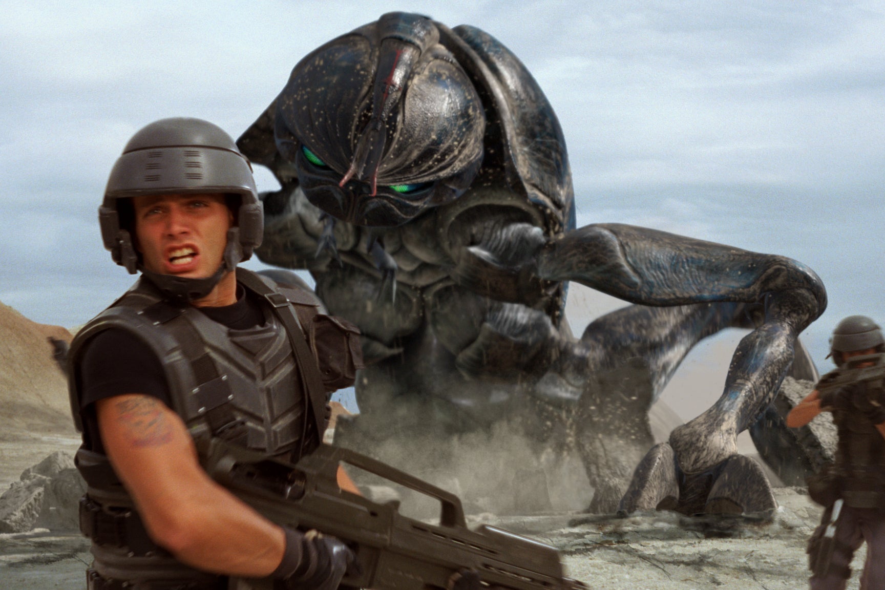 A still of Casper Van Dien in the foreground wearing tactical gear and armor, holding a gun, while running from a large alien in the background. 