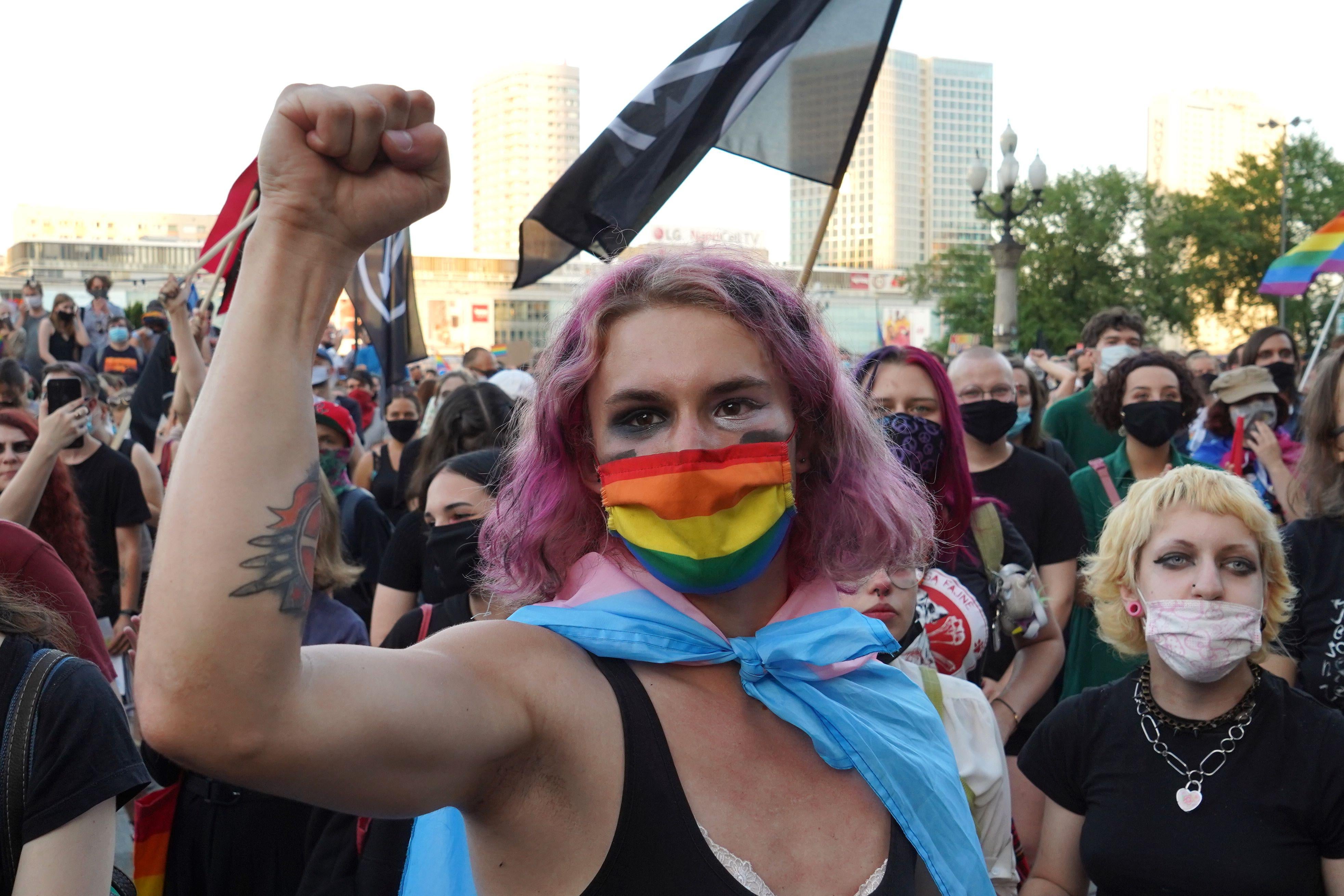 Activist with pink hair holds up her fist in front of a rainbow flag. 