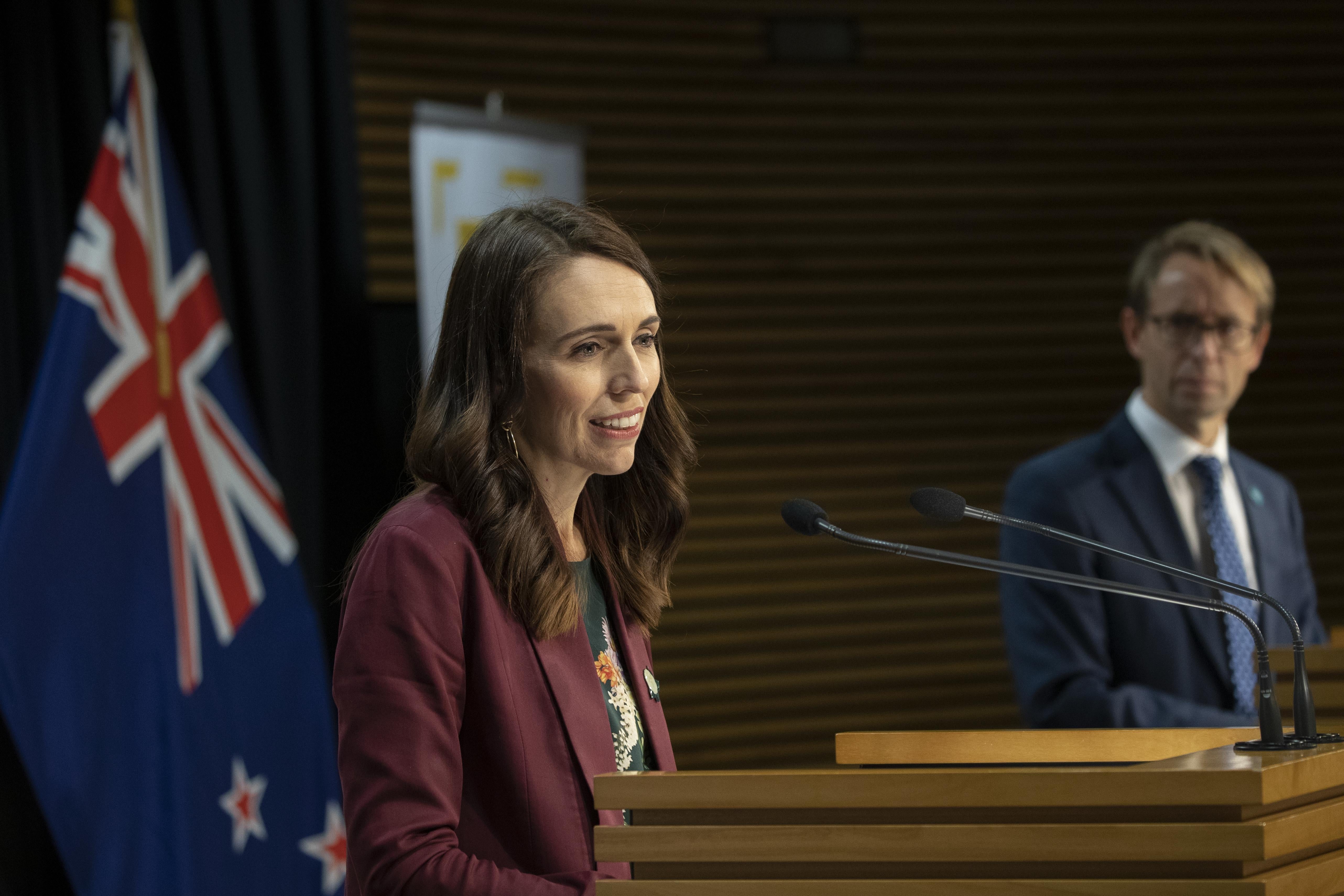Jacinda Ardern speaks at a podium. Behind her is the New Zealand flag.