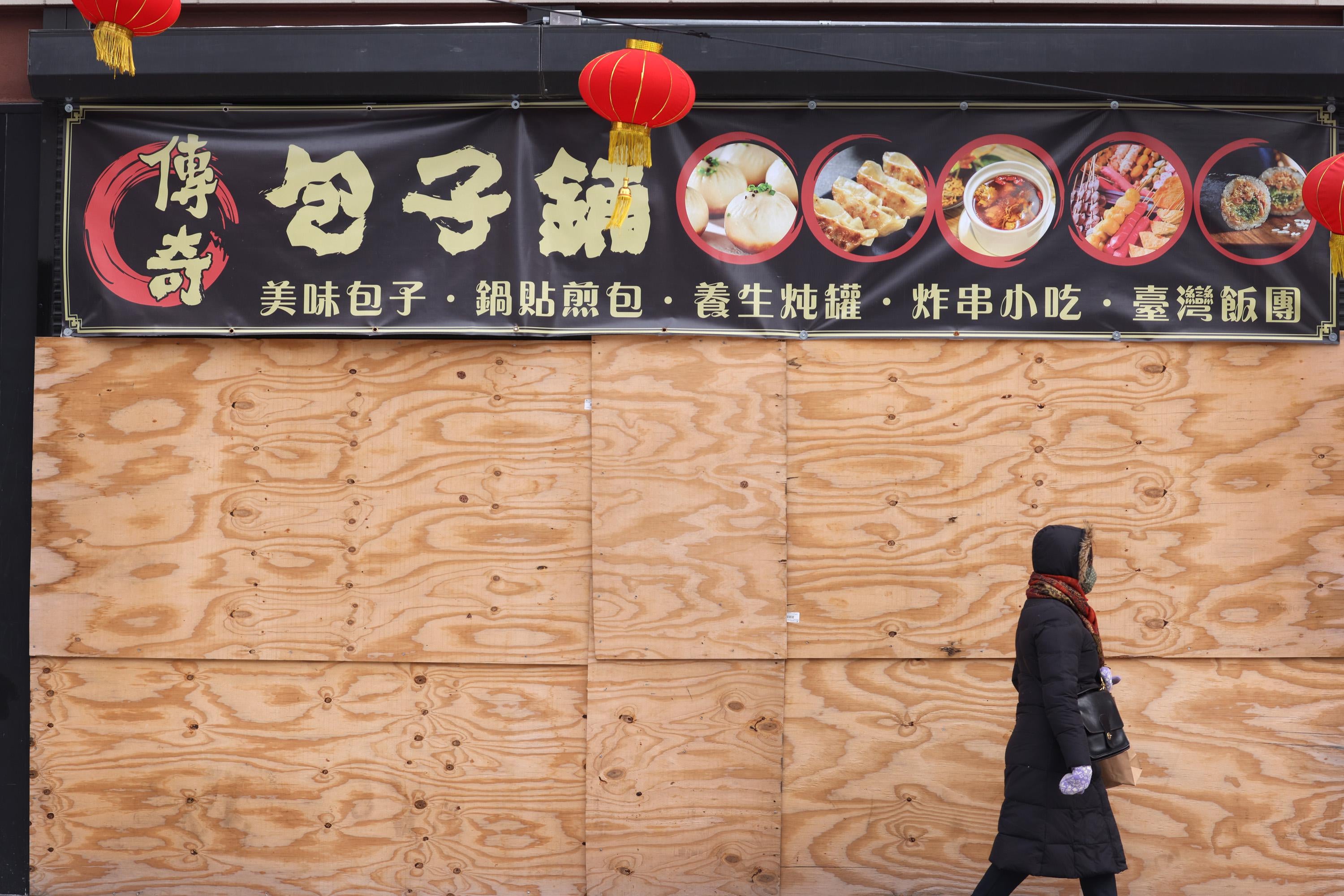 A person in a coat walks past a boarded-up restaurant.