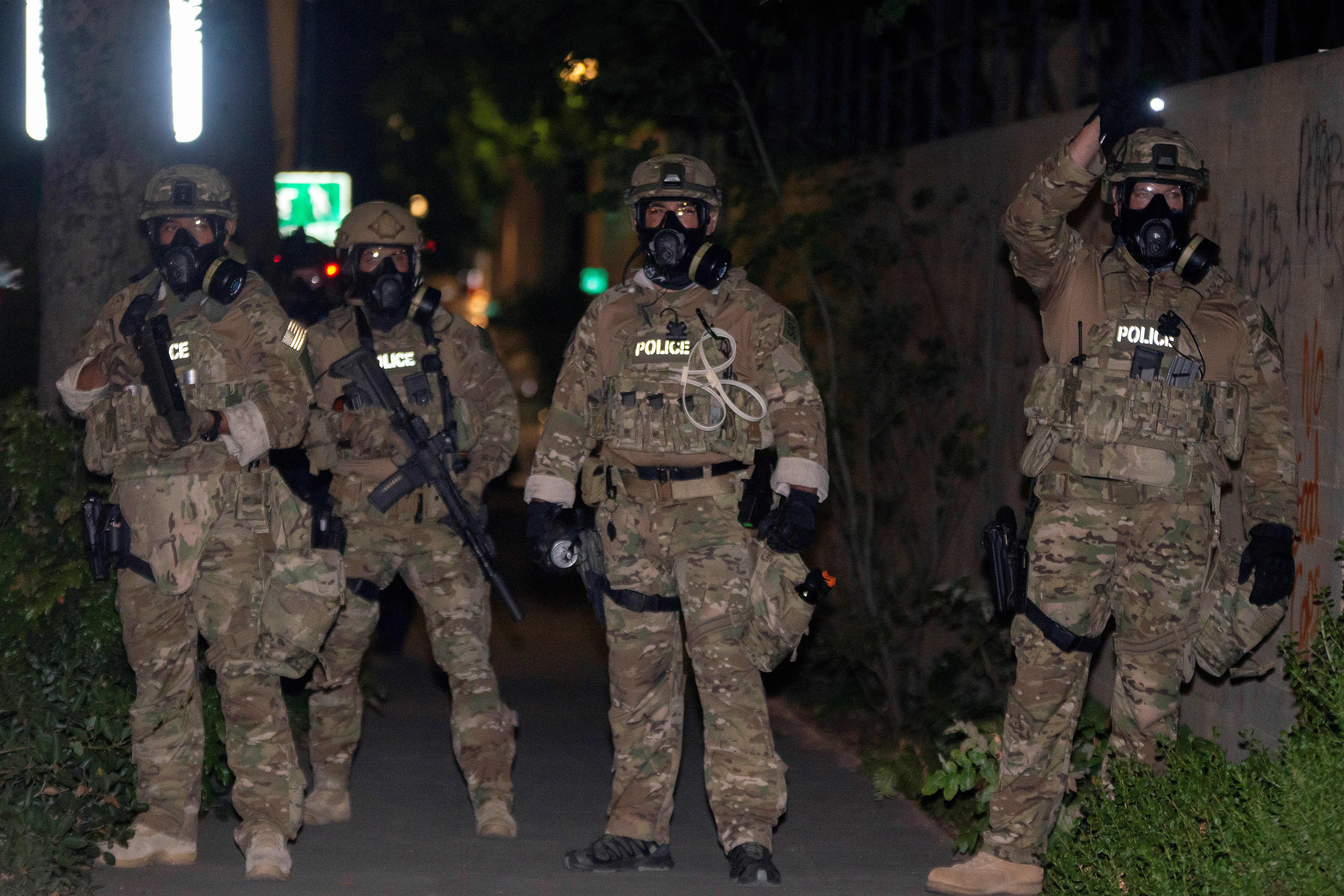 Four federal law enforcement officers wearing gas masks and full-body camo with the word POLICE emblazoned on the chest, at night. Two are holding guns.
