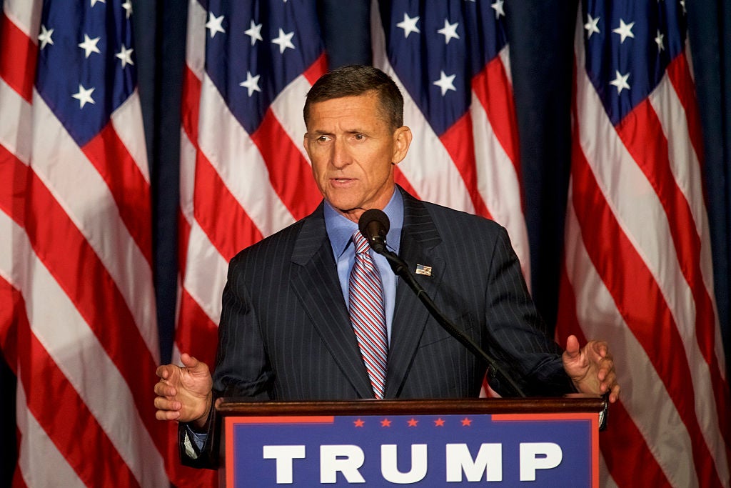 Flynn, wearing a pinstriped suit, speaks from behind a TRUMP logo on a lectern and in front of a row of American flags.
