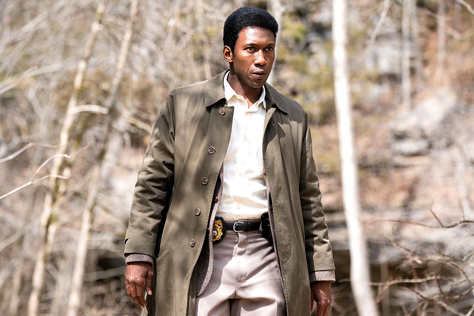 Mahershala Ali as Detective Wayne Hays, wearing a trenchcoat and walking in the woods.