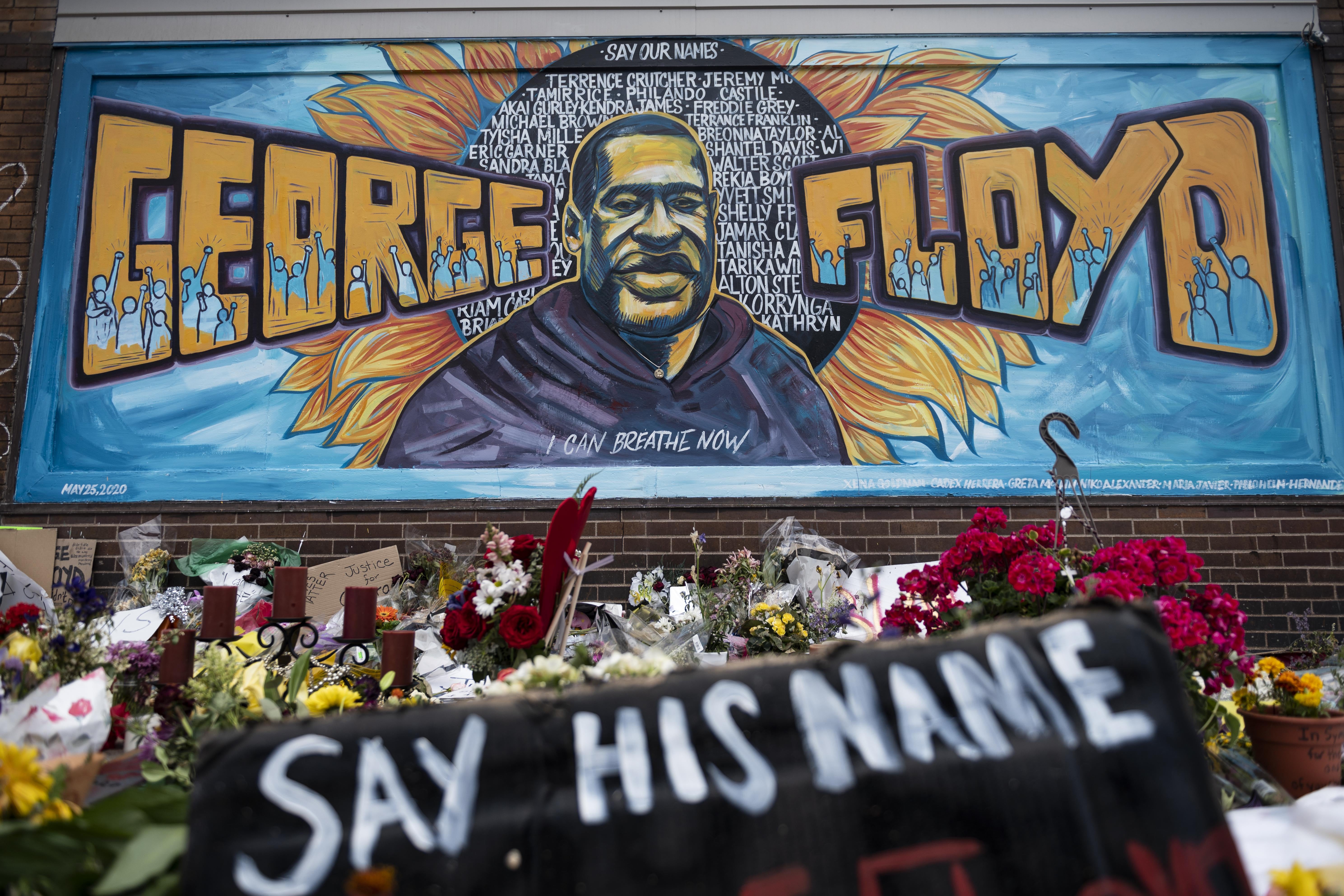 A mural with George Floyd's face and name is seen. Flowers stand below it along with a spray-painted cloth that says "Say His Name."