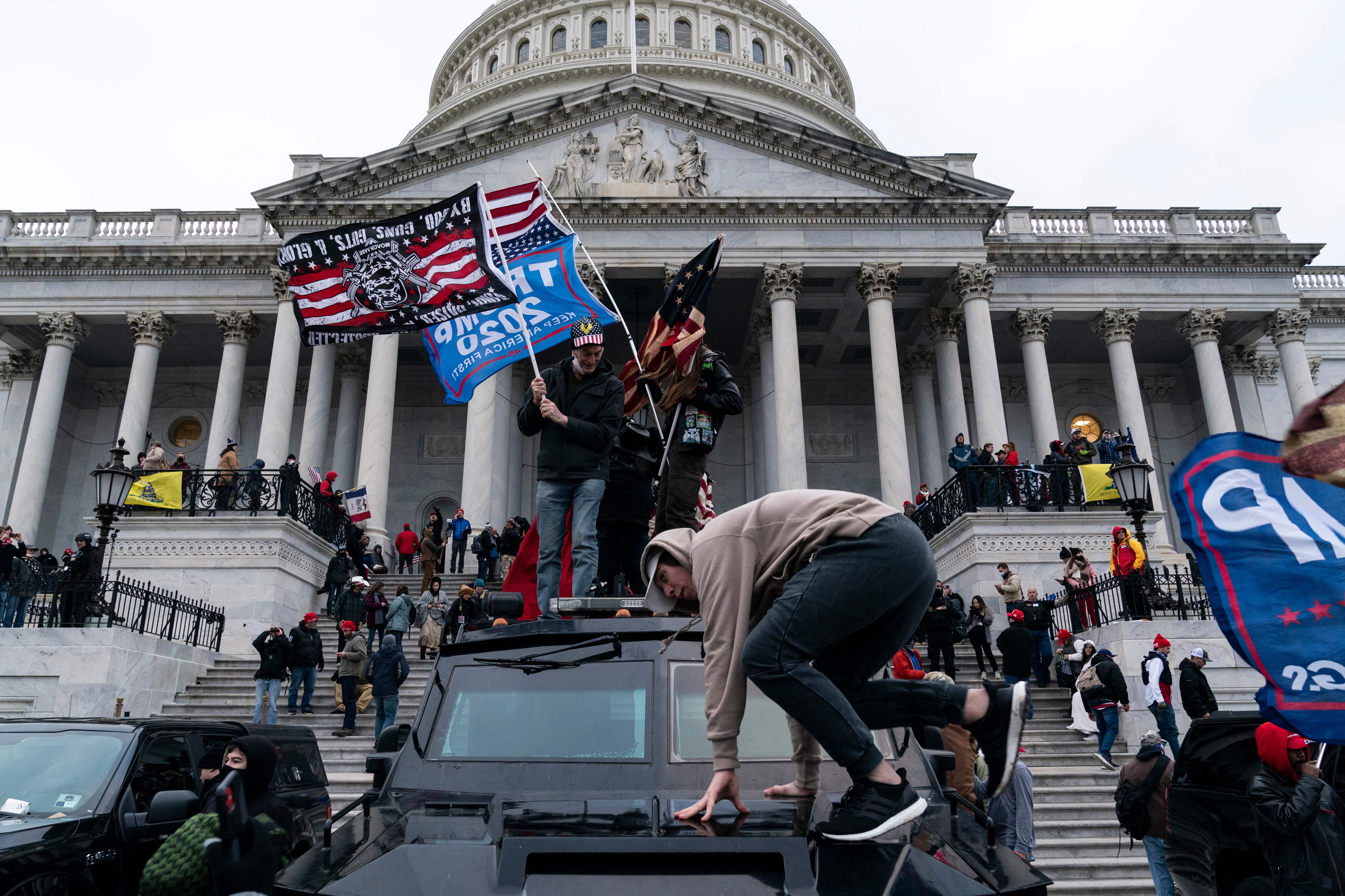 A man jumps on a vehicle in front of the Capitol. Others stand on top of it, waving large "Trump 2020" flags. More protesters are massed on the Capitol steps.