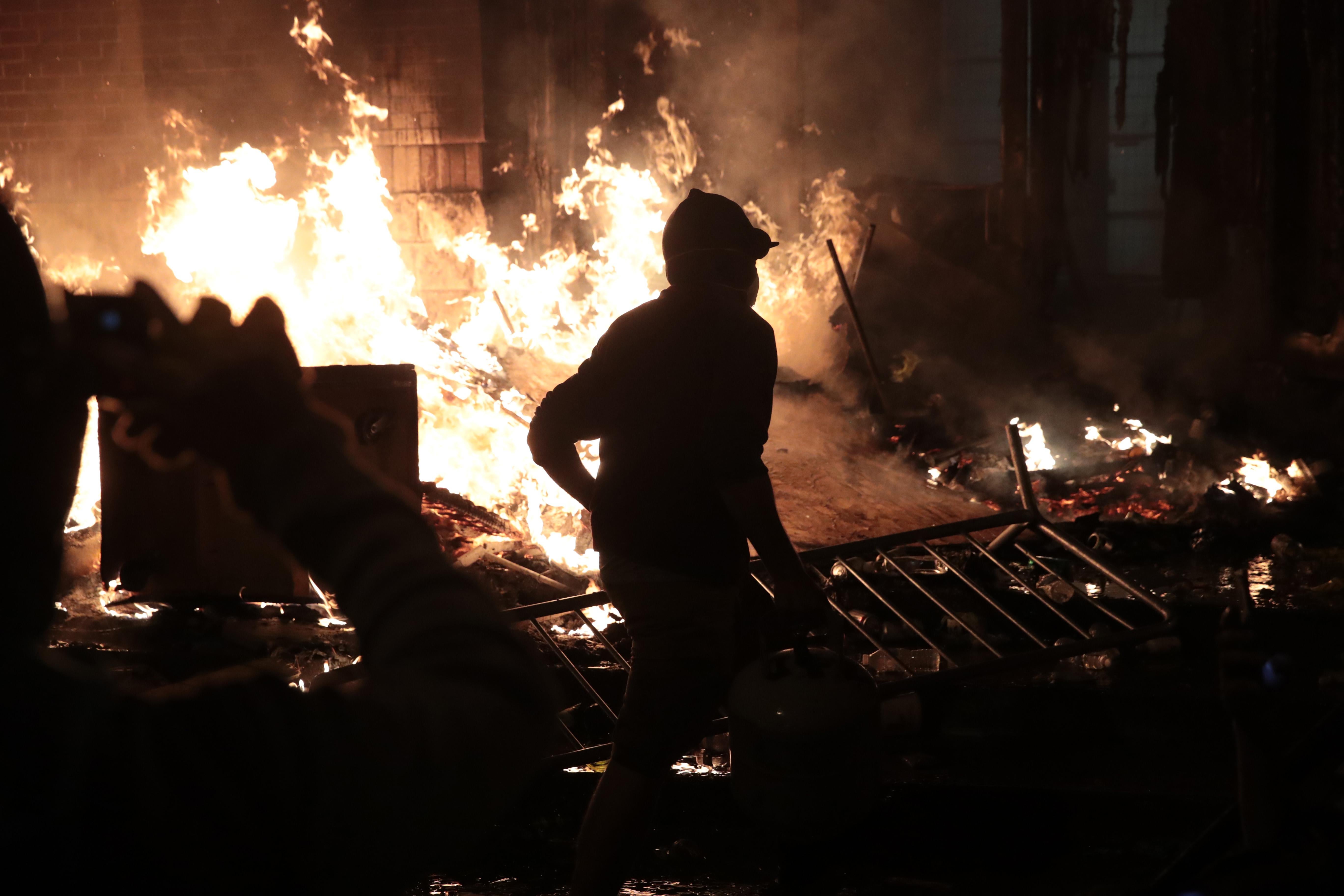 A silhouetted protester stands in front of bright flames while another person films them and a fallen metal crowd barrier reflects the light. 