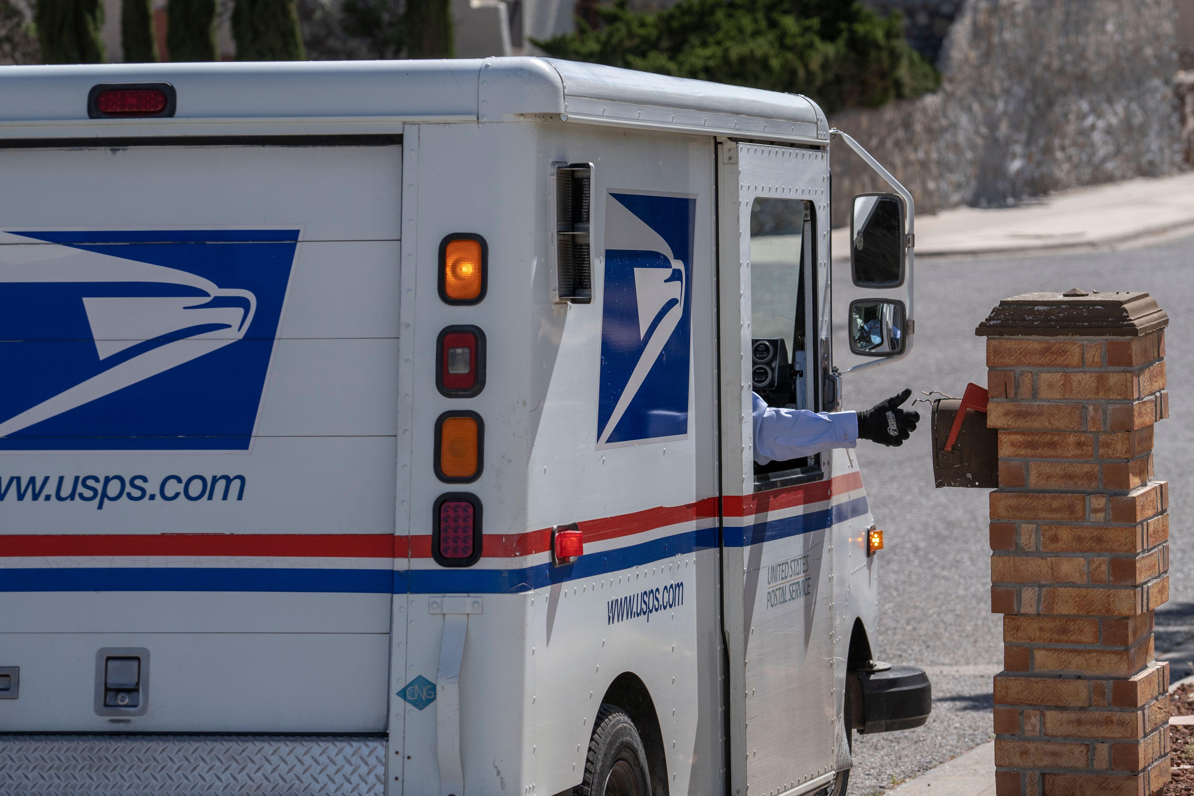 A hand sticks out the window of a mail carrier to place mail inside a mailbox.