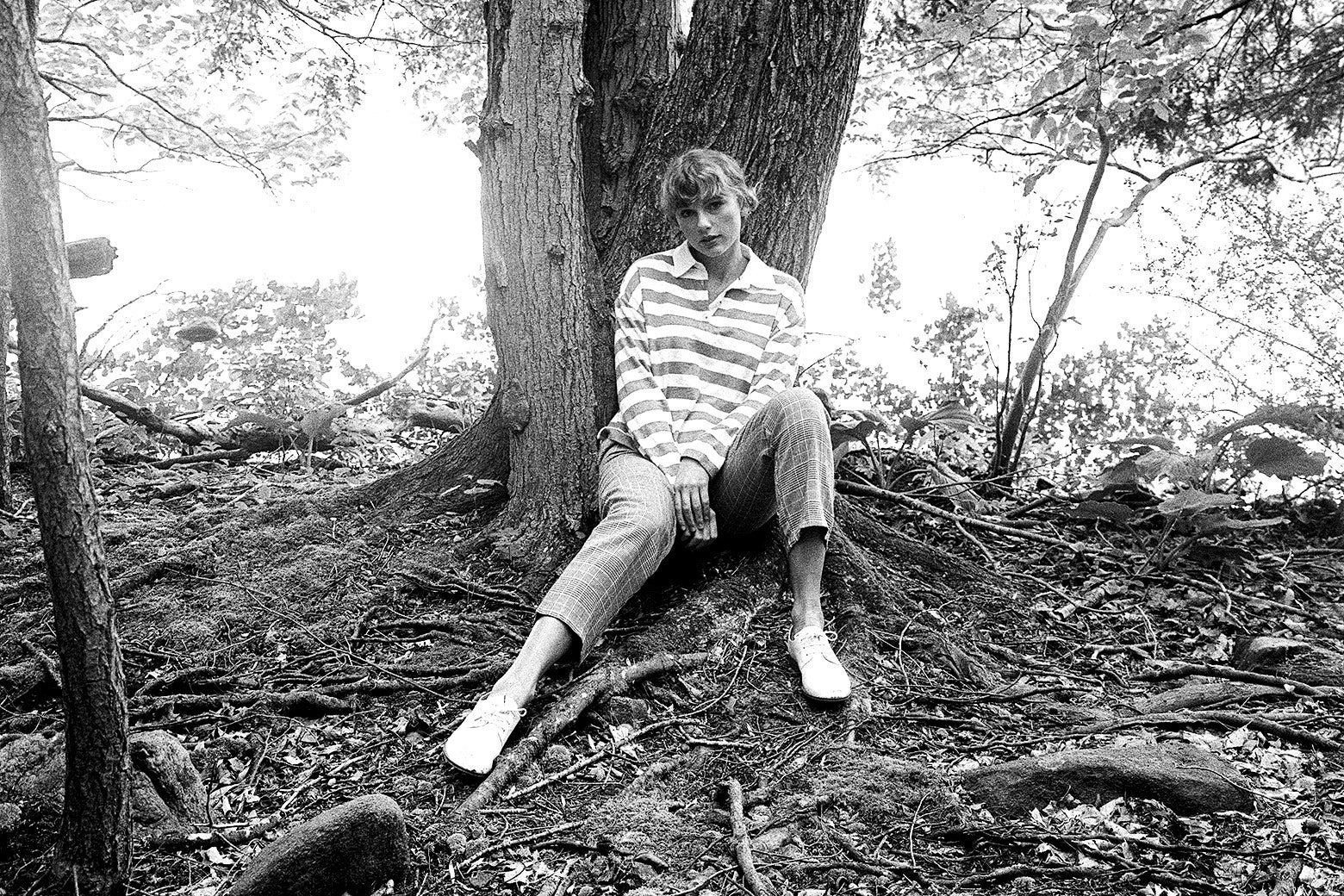 Taylor Swift sits on the ground in front of a tree.