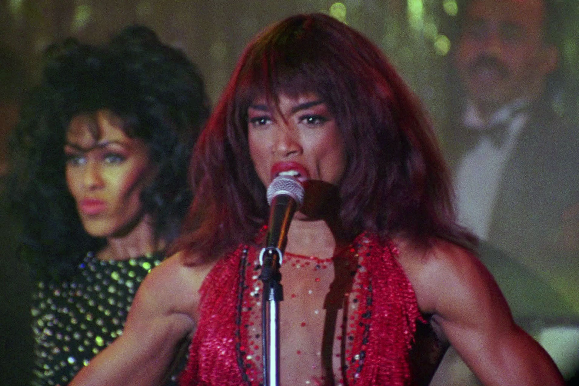 Angela Bassett stands on-stage with her arms outstretched, mid song,  dressed as Tina Turner with long hair and a sparkly red costume on and a background singer behind her.