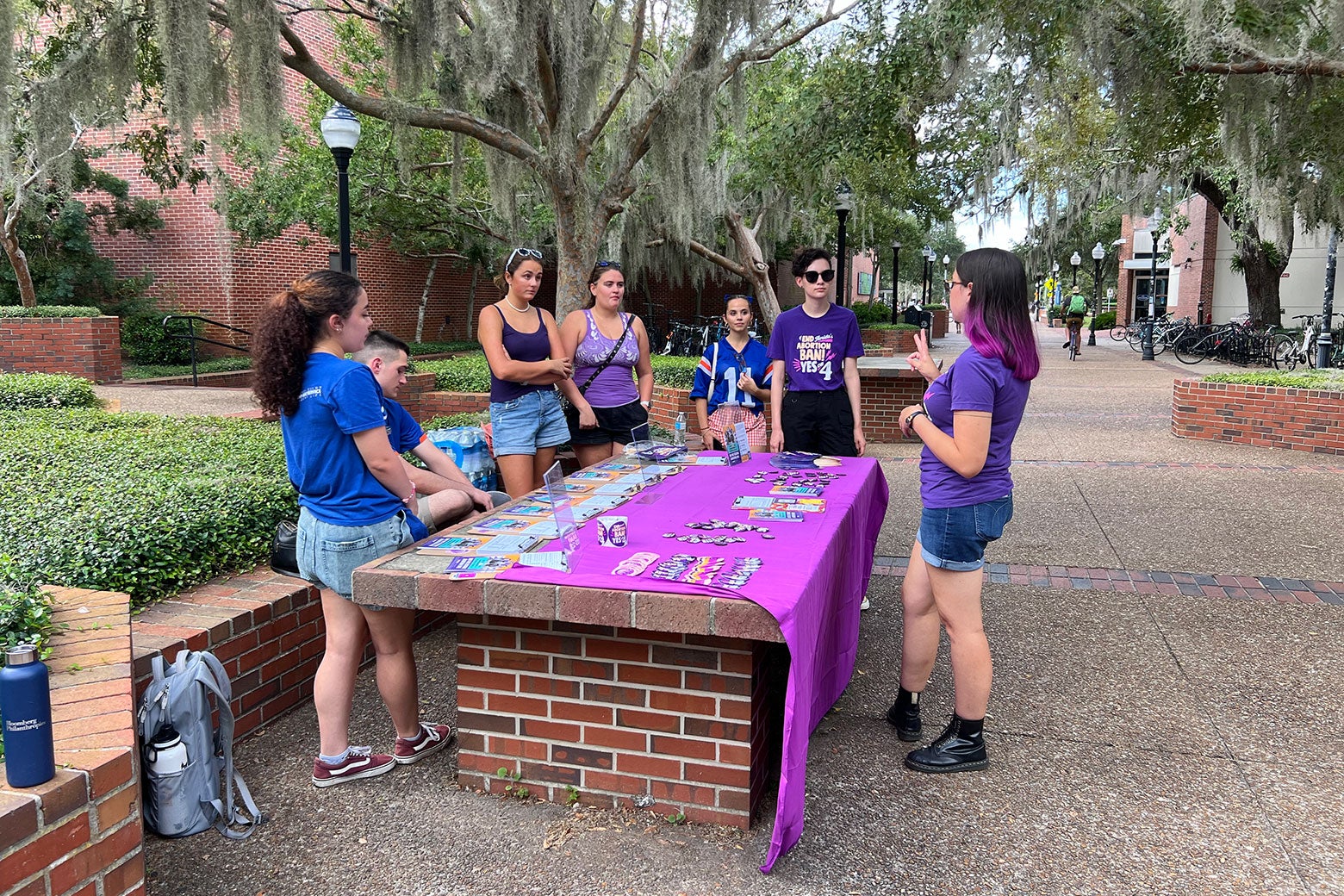 A young woman in a purple T-shirt with purple hair talks to other students standing around a table on a college campus.