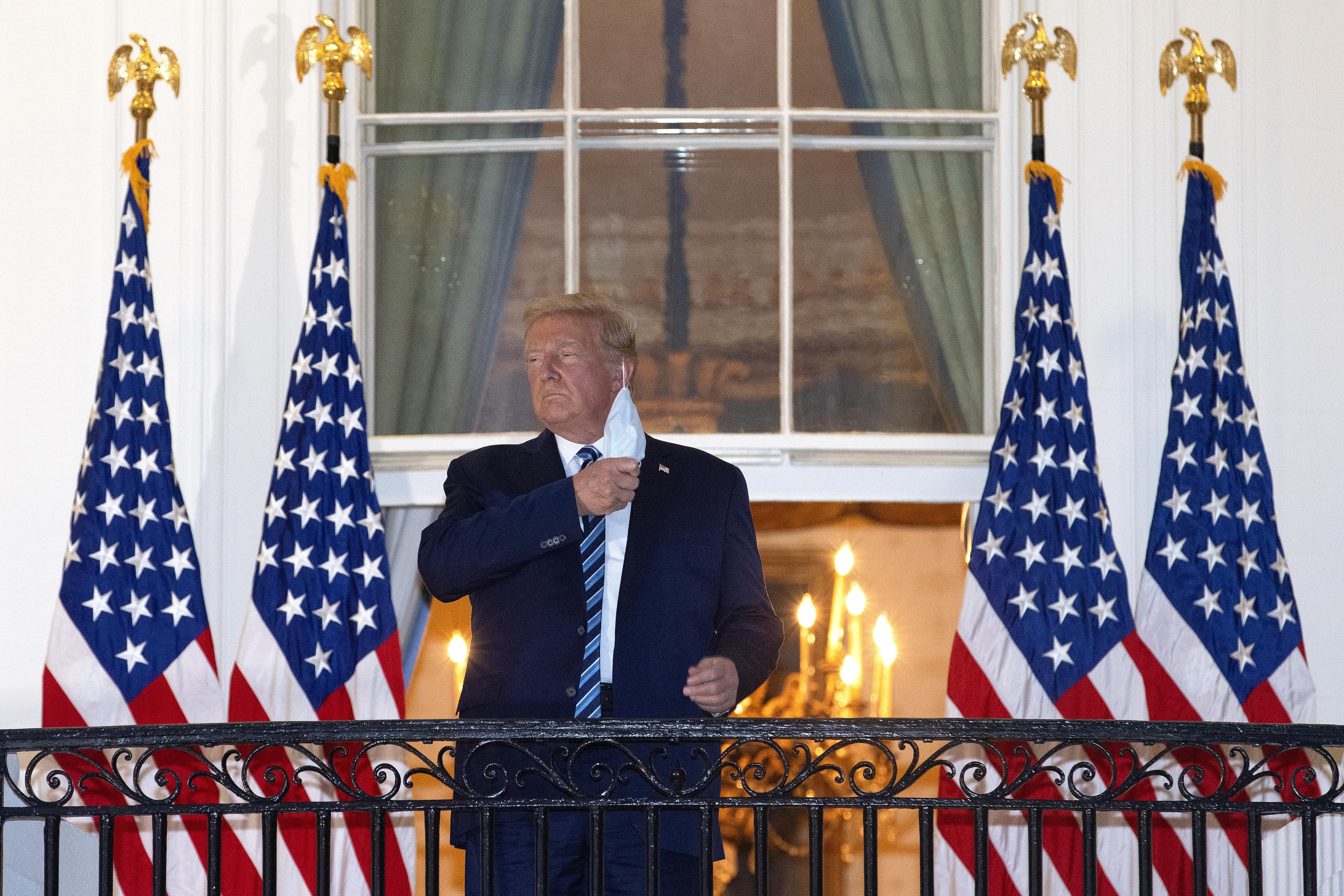 Donald Trump removes his mask while standing on a balcony with U.S. flags