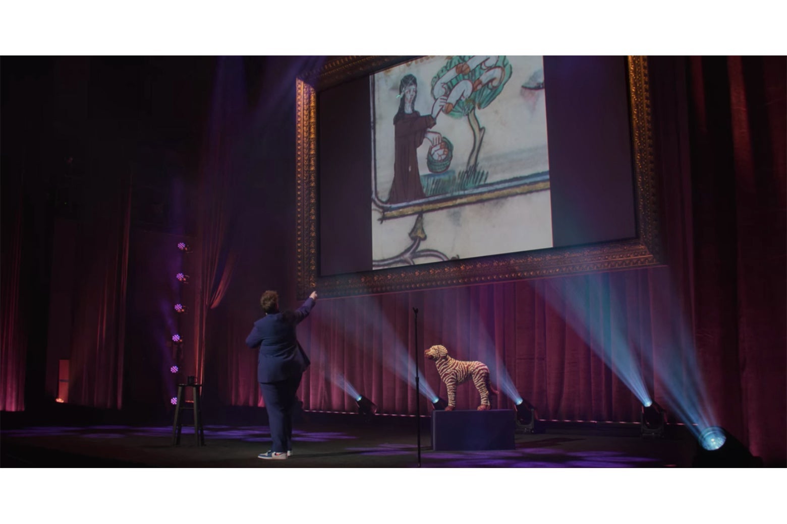 Hannah Gadsby onstage in front of a detail from an illuminated manuscript showing a nun picking penises from a penis tree.