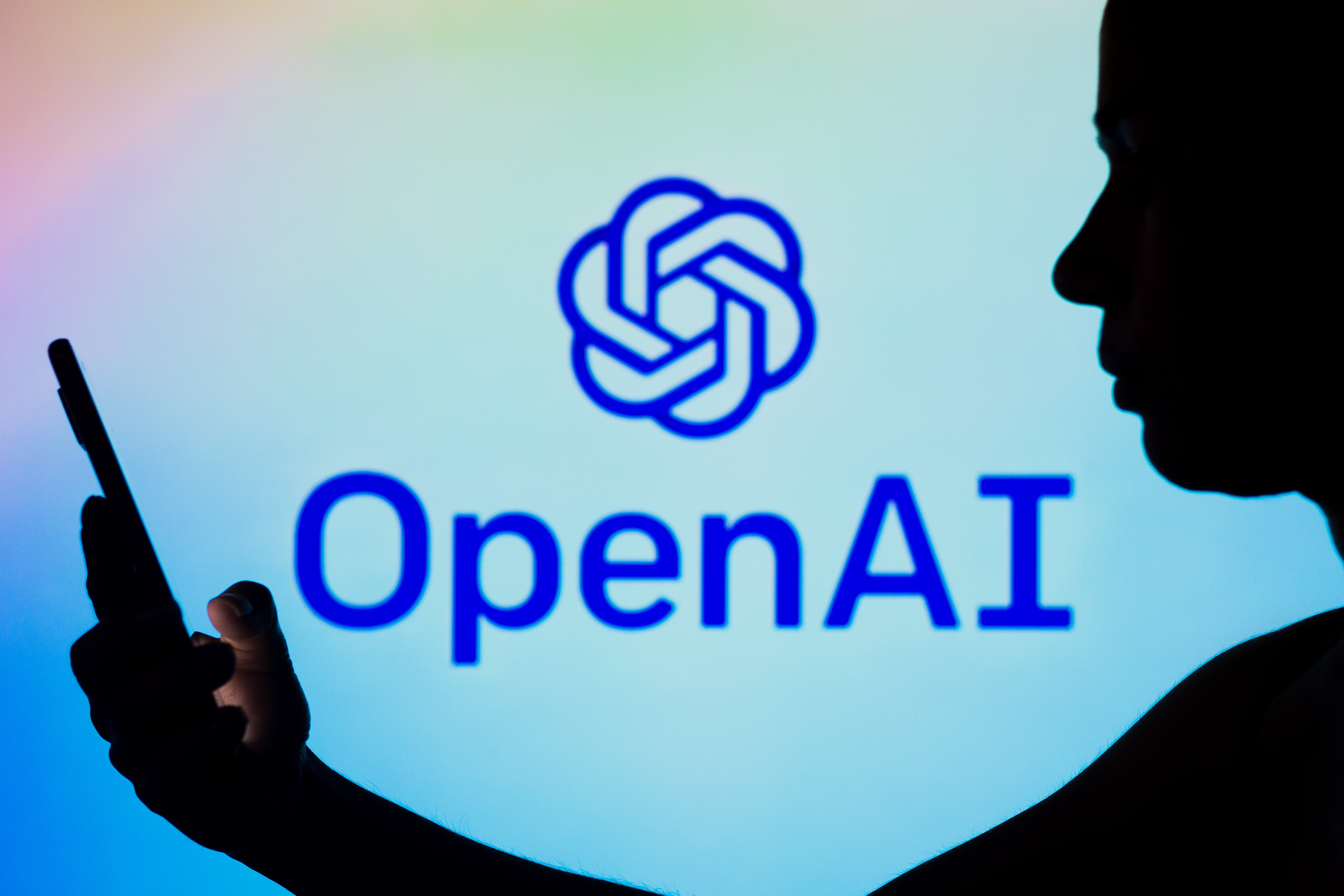 A silhouetted person talked to a smartphone, against the OpenAI logo.