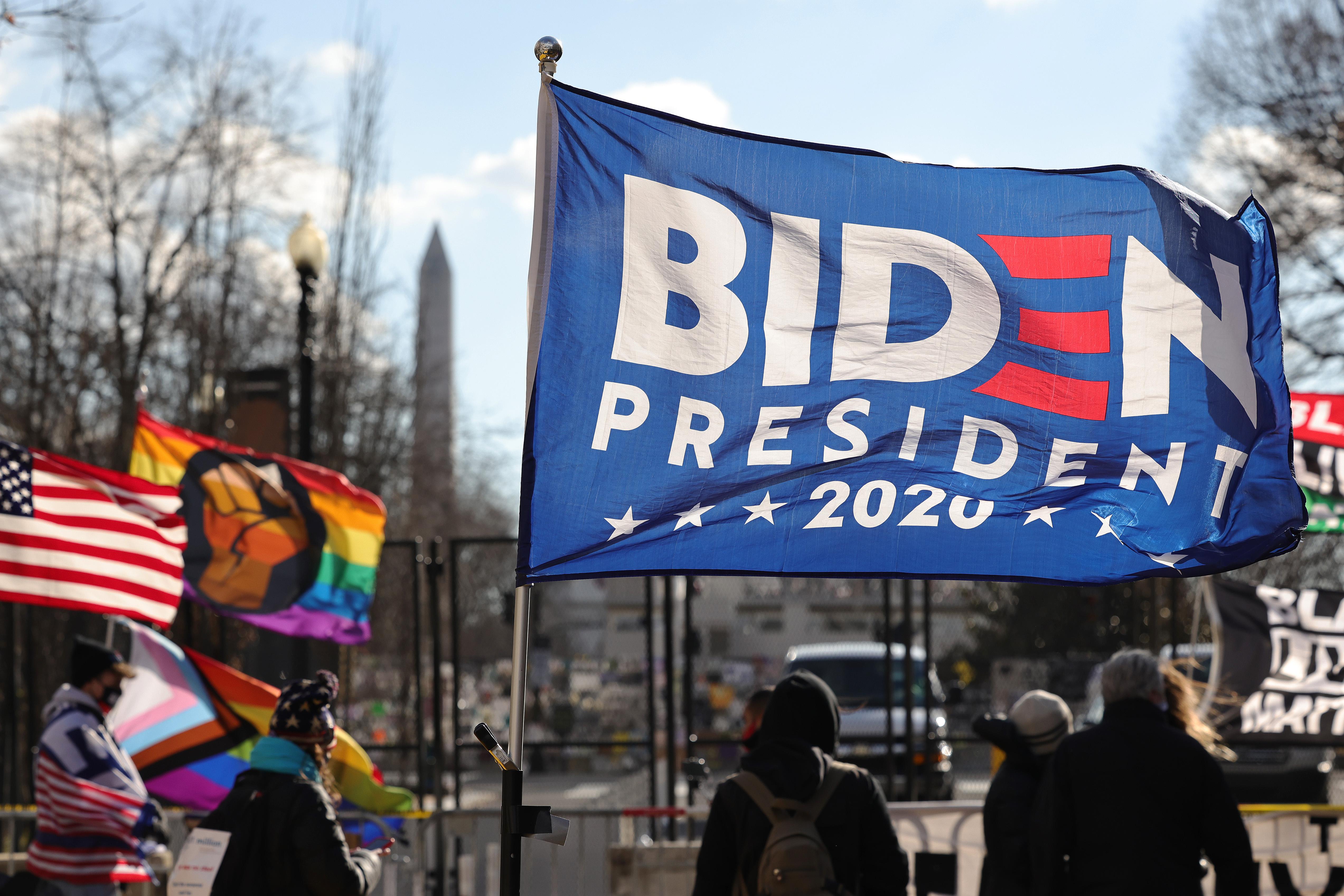 A Biden flag waves in the wind, with rainbow flags in the background