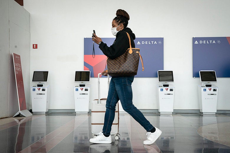 A woman wearing a face mask and looking at her cellphone walks through an airport.