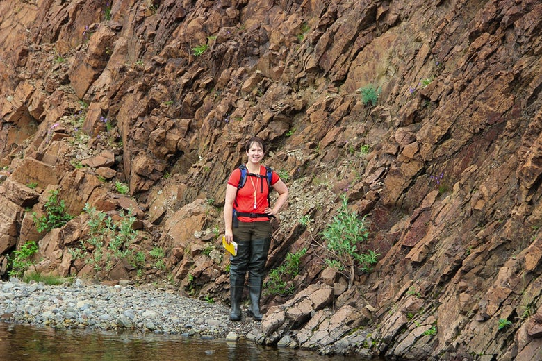 Woman wearing a red shirt standing next to a rocky cliff face near a stream of water.